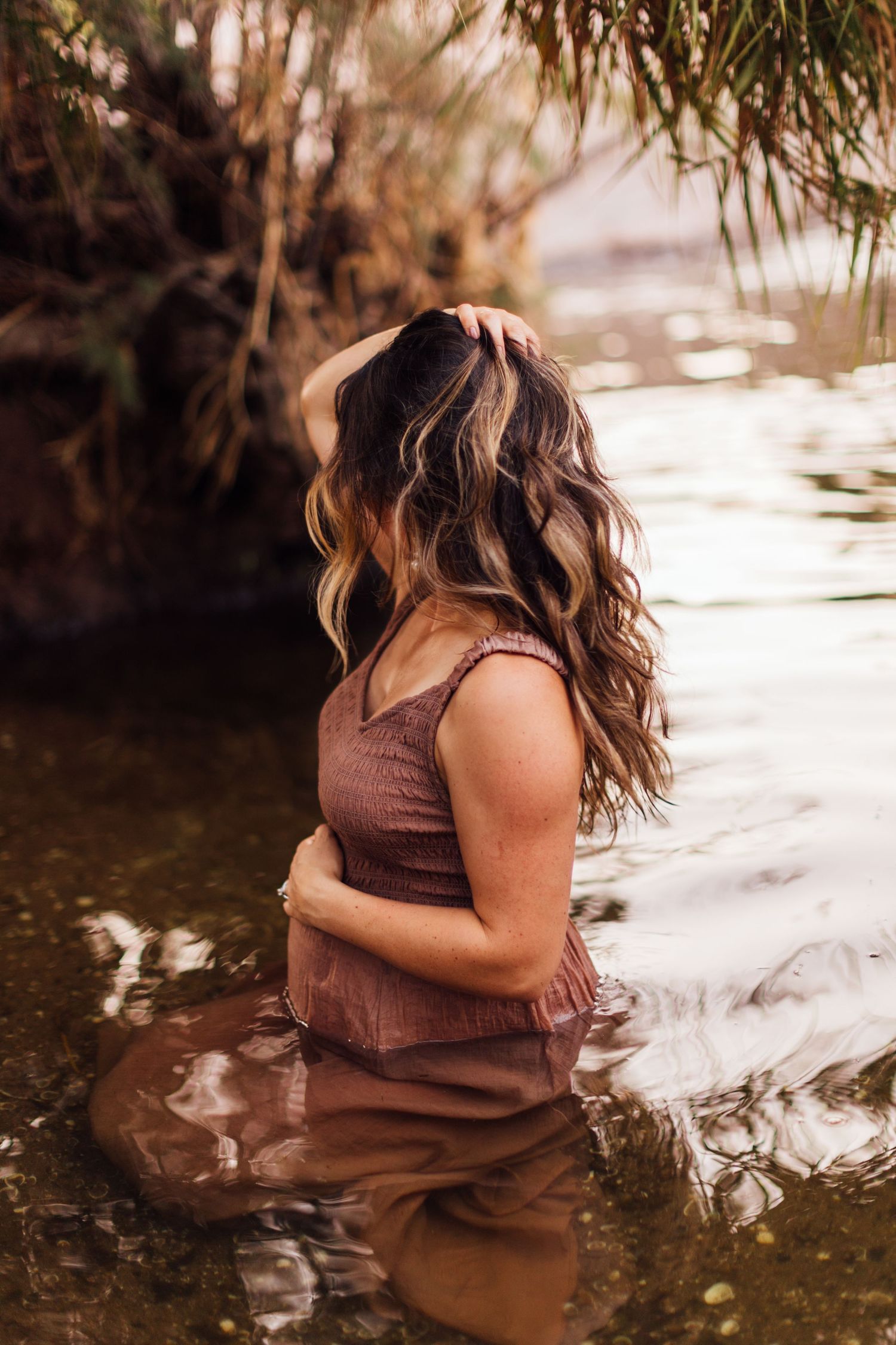 Woman in brown dress standing in shallow water with long wavy hair, surrounded by lush greenery.