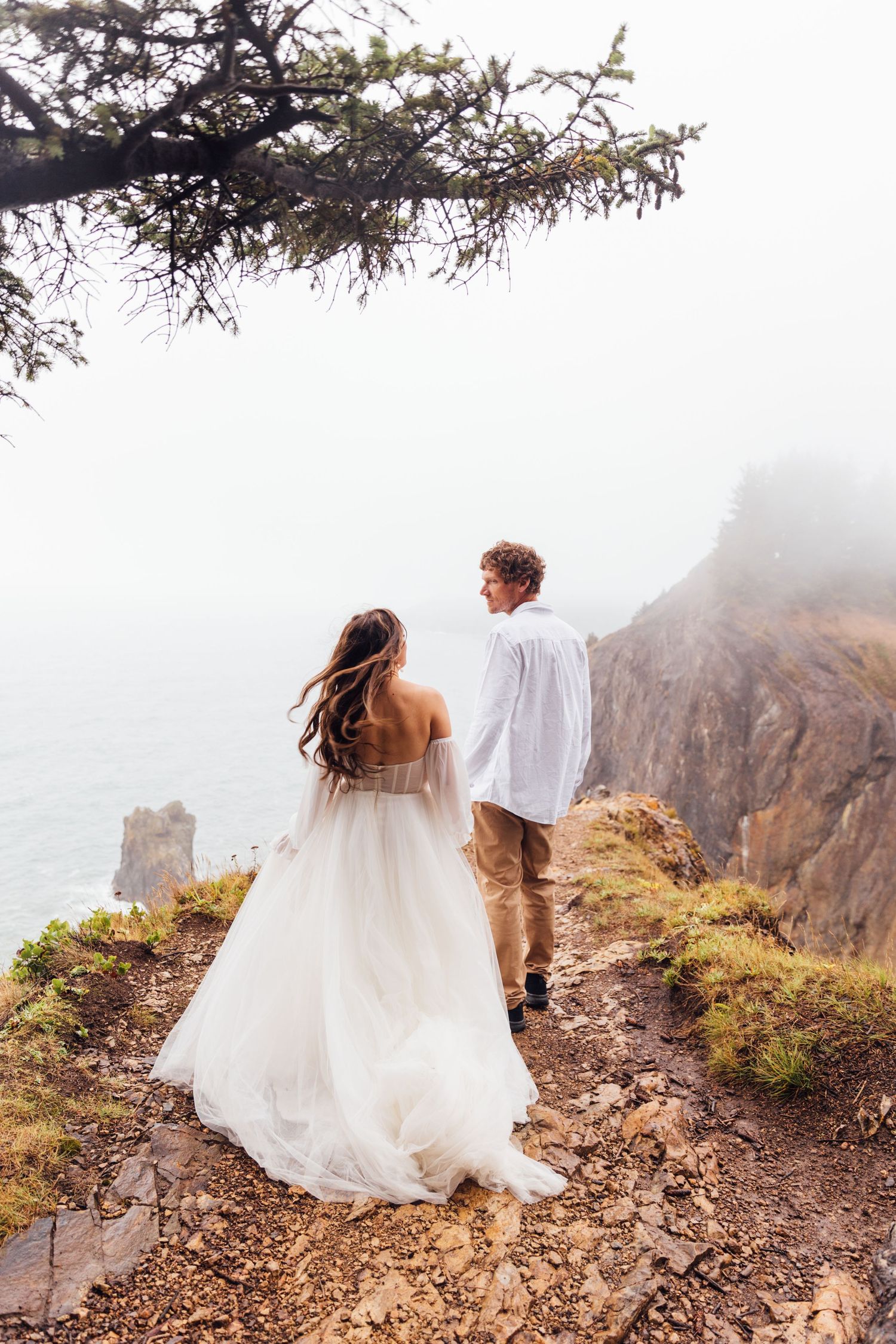 Bride in white gown and groom in white shirt stand on misty coastal cliff, holding hands with ocean views.