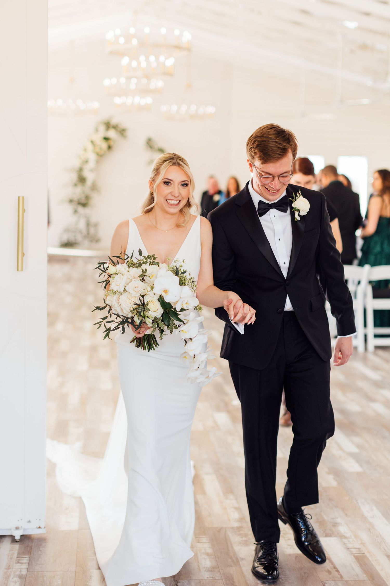 Wedding couple walks together in bright white venue decorated with elegant greenery.