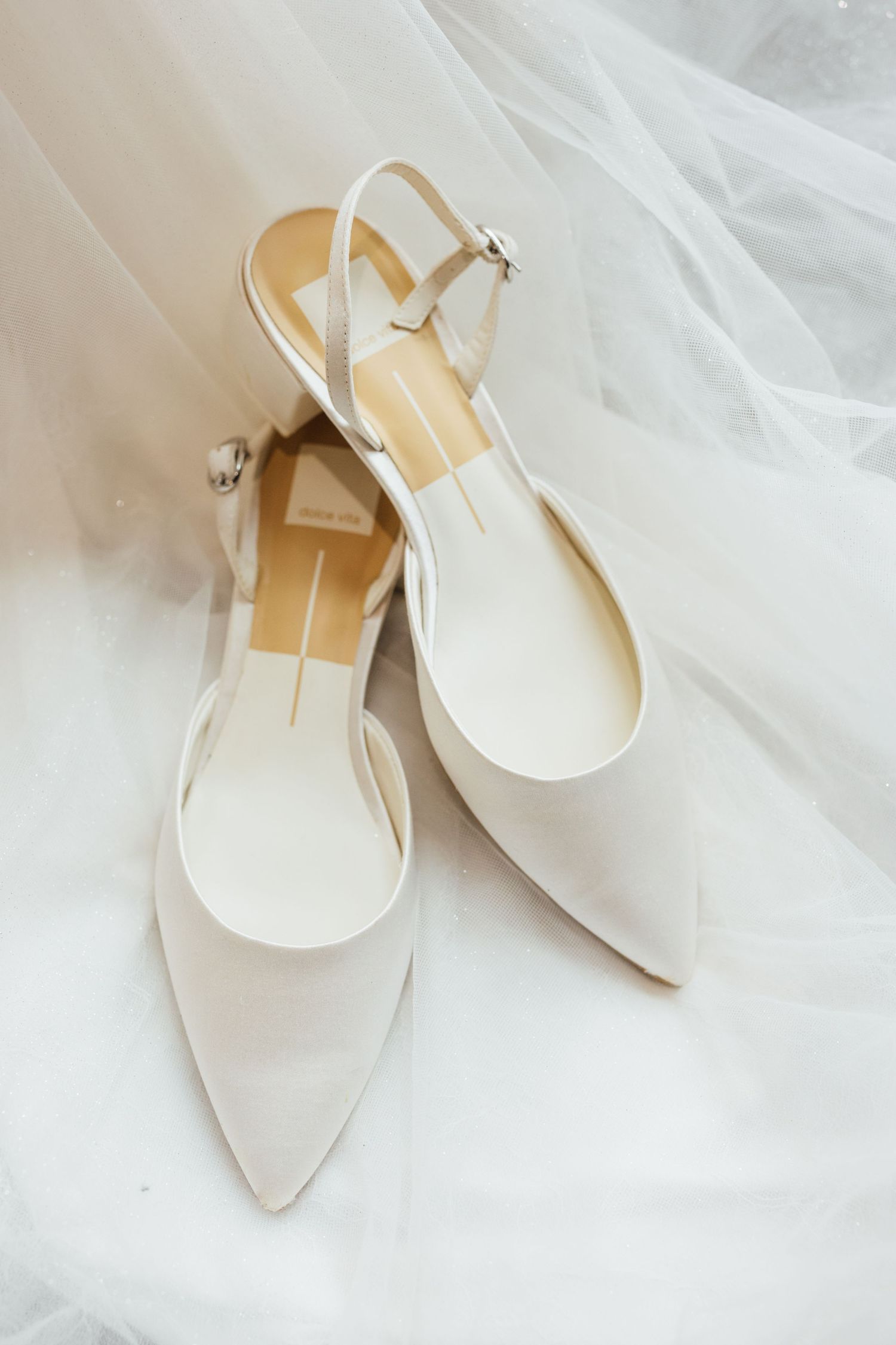 Elegant white slingback kitten heel shoes with pointed toes displayed on soft white fabric background.