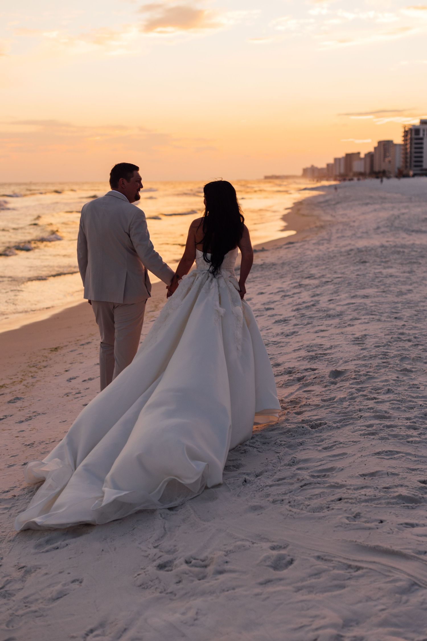 Bride and groom holding hands walking on a sandy beach at sunset, with waves and city skyline in the background.
