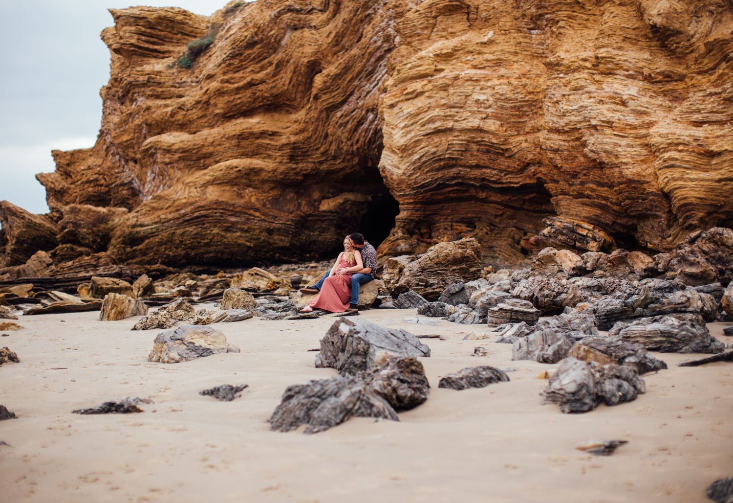 Person in colorful dress sitting among rocks beside a large sandstone cliff on a sandy beach.
