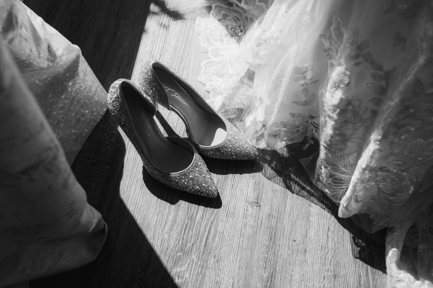 A pair of sparkly high heel bridal shoes resting on a wooden floor next to white fabric.