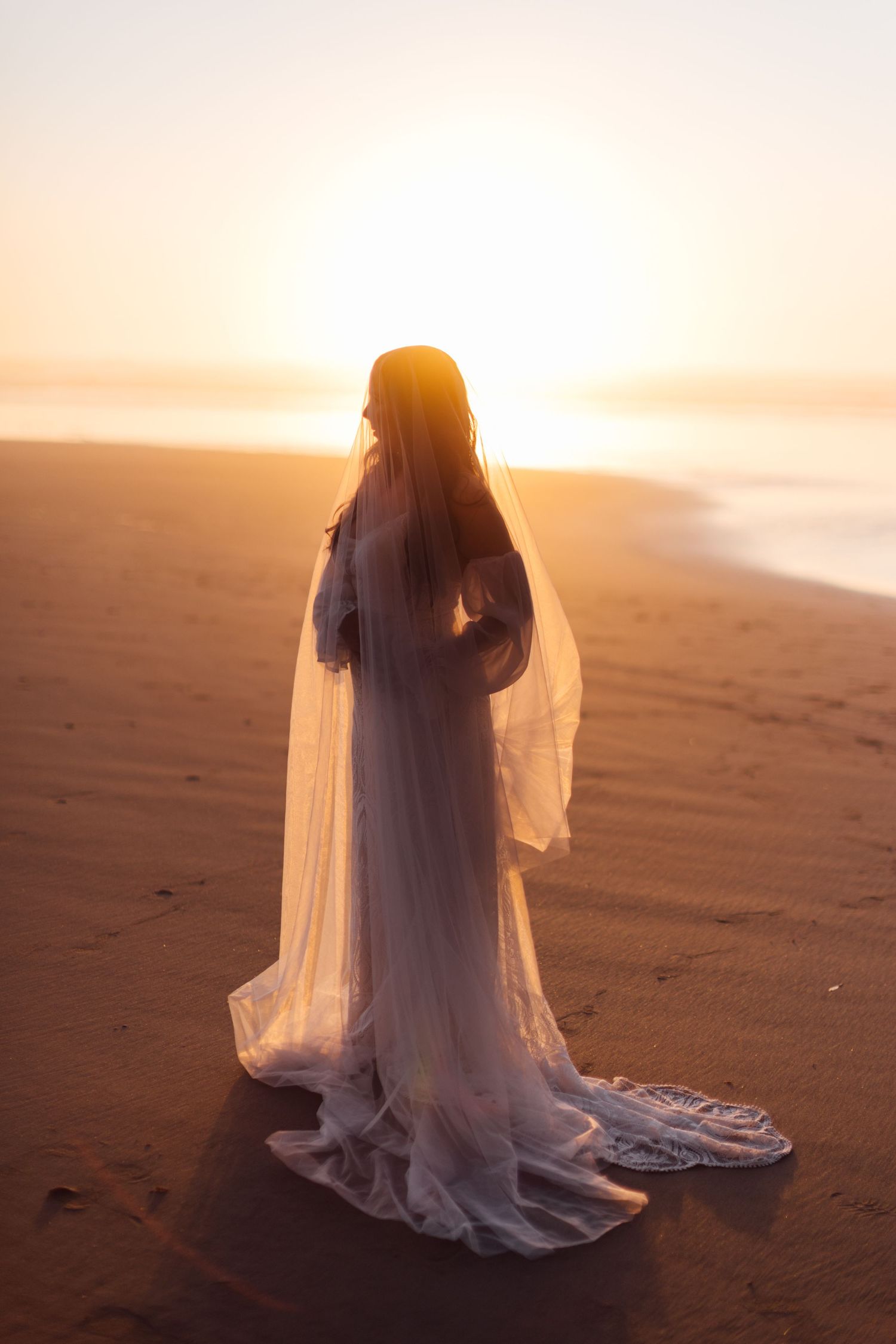 A bride in a flowing white gown and veil stands on a sandy beach at golden sunset.