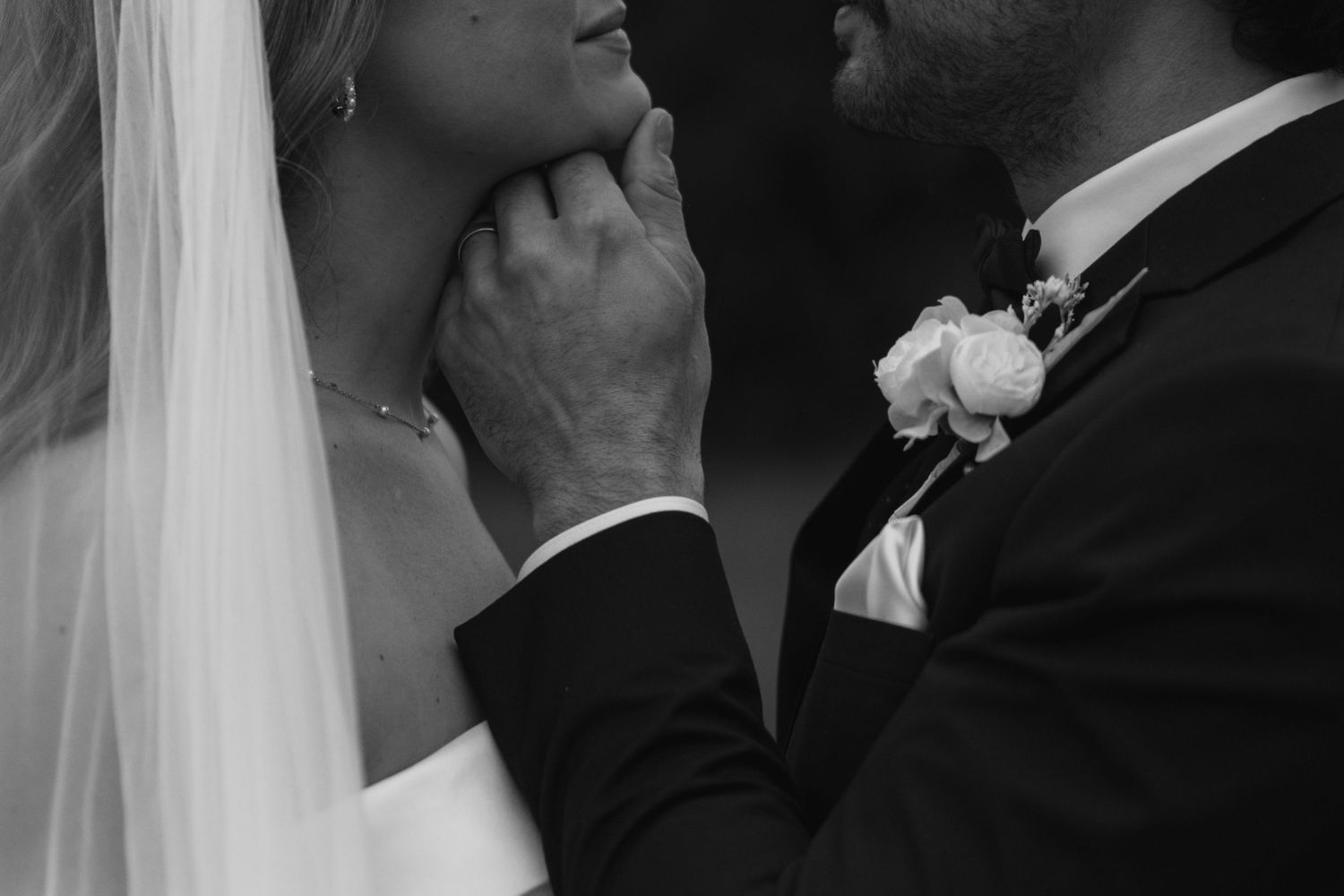 Black and white wedding photo of groom tenderly holding brides chin, both in elegant attire with floral boutonniere.