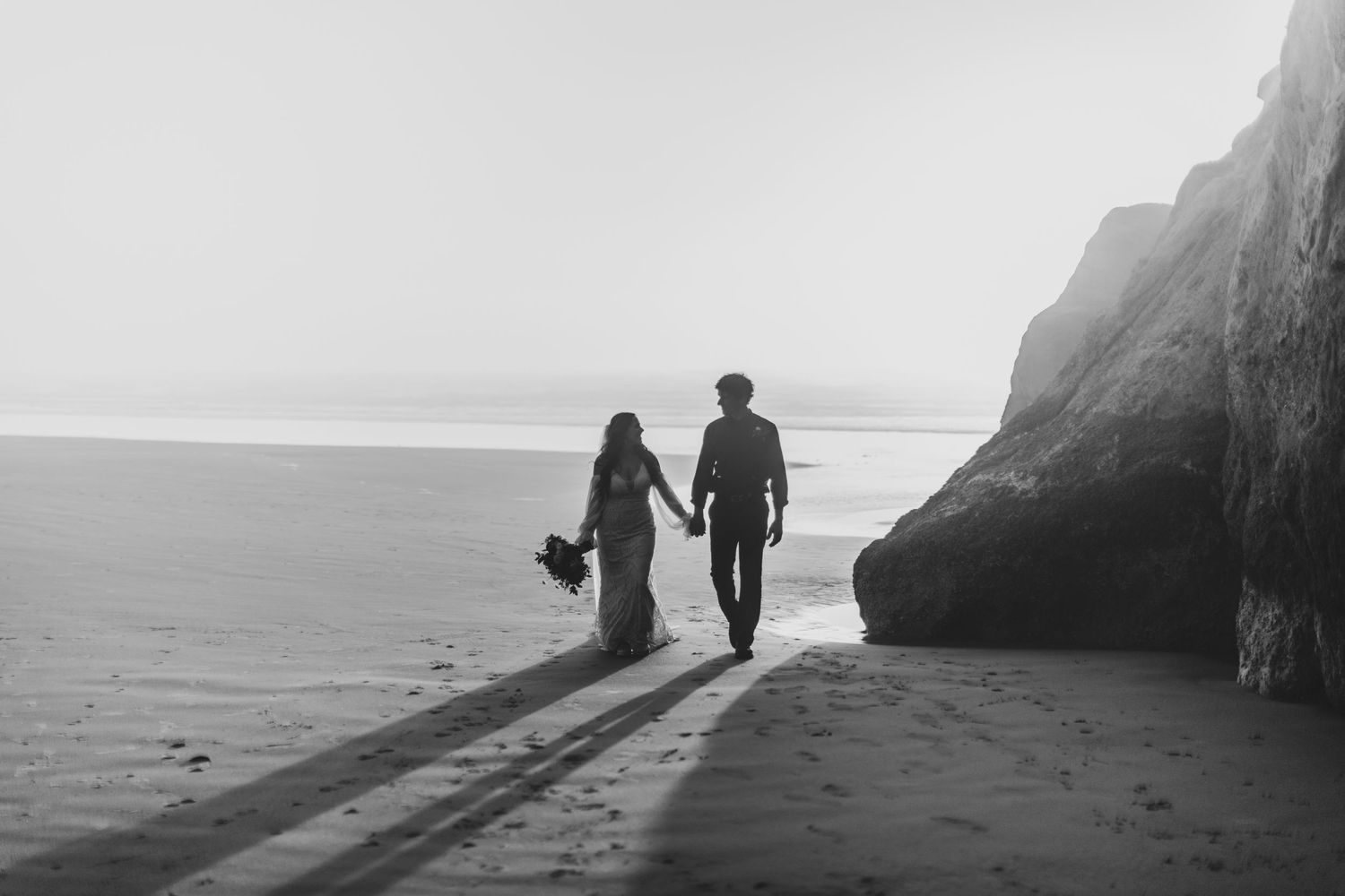 Black and white photo of a couple holding hands walking on a beach near rocky cliffs toward the ocean.