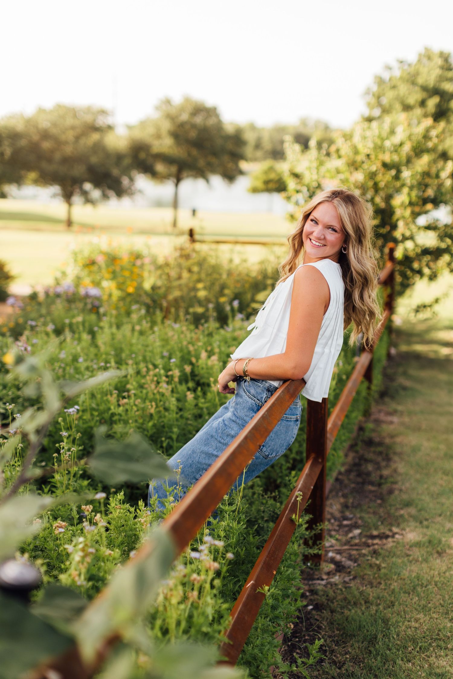 Young woman in white top and jeans smiling by a wooden fence surrounded by a lush garden at golden hour.
