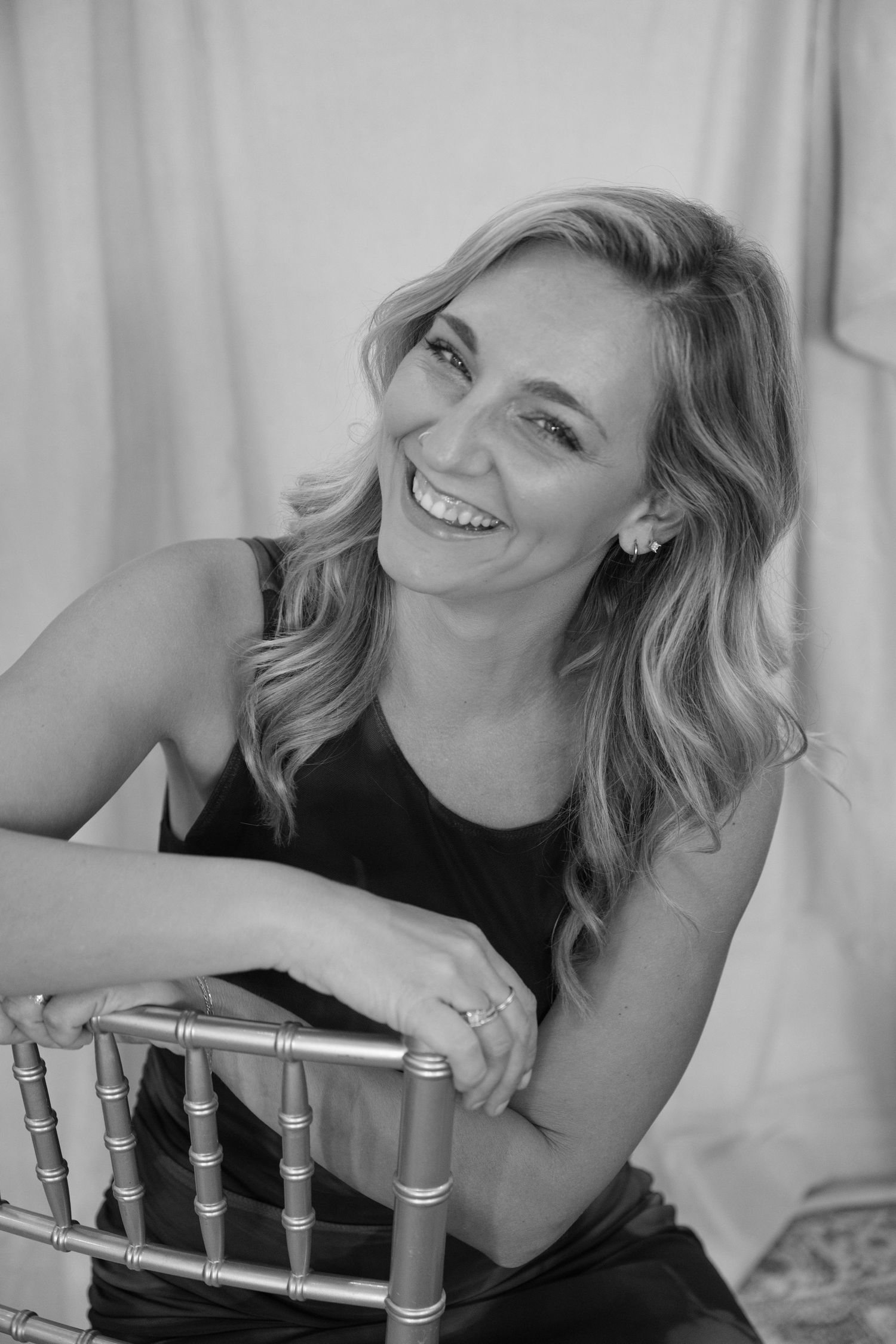 Smiling blonde woman laughing joyfully while leaning on a gold chiavari chair in a black and white portrait photo.