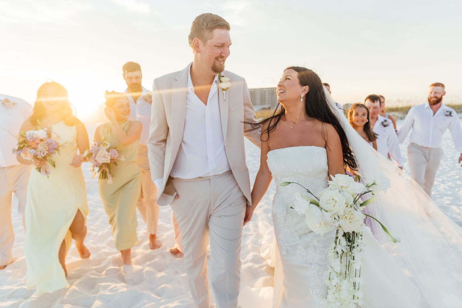 Bride and groom walk hand-in-hand on a sunny beach with wedding party, bride holding white floral bouquet.