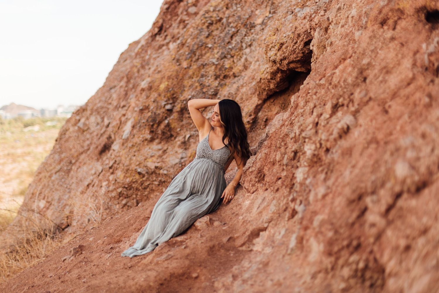 Woman in gray dress poses gracefully against red rock formation in arid landscape.