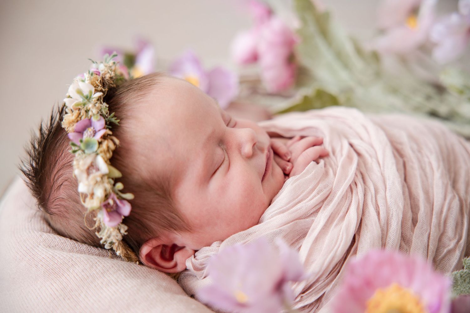 Newborn baby sleeping peacefully wearing a delicate floral crown and pink wrap in soft natural light.