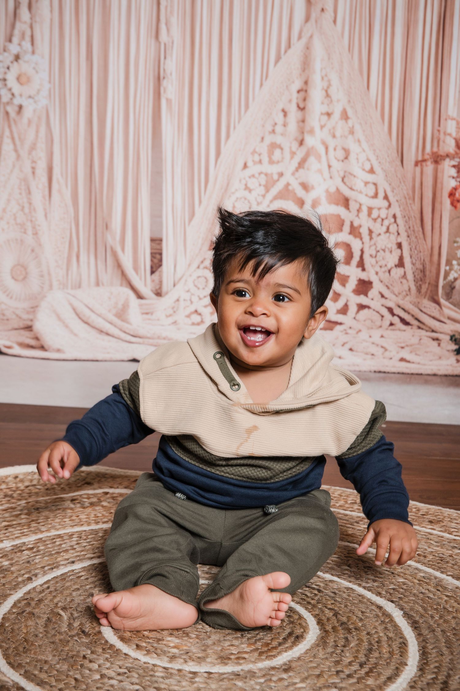 Happy young child sits on circular patterned carpet wearing beige scarf and olive pants against decorative backdrop.