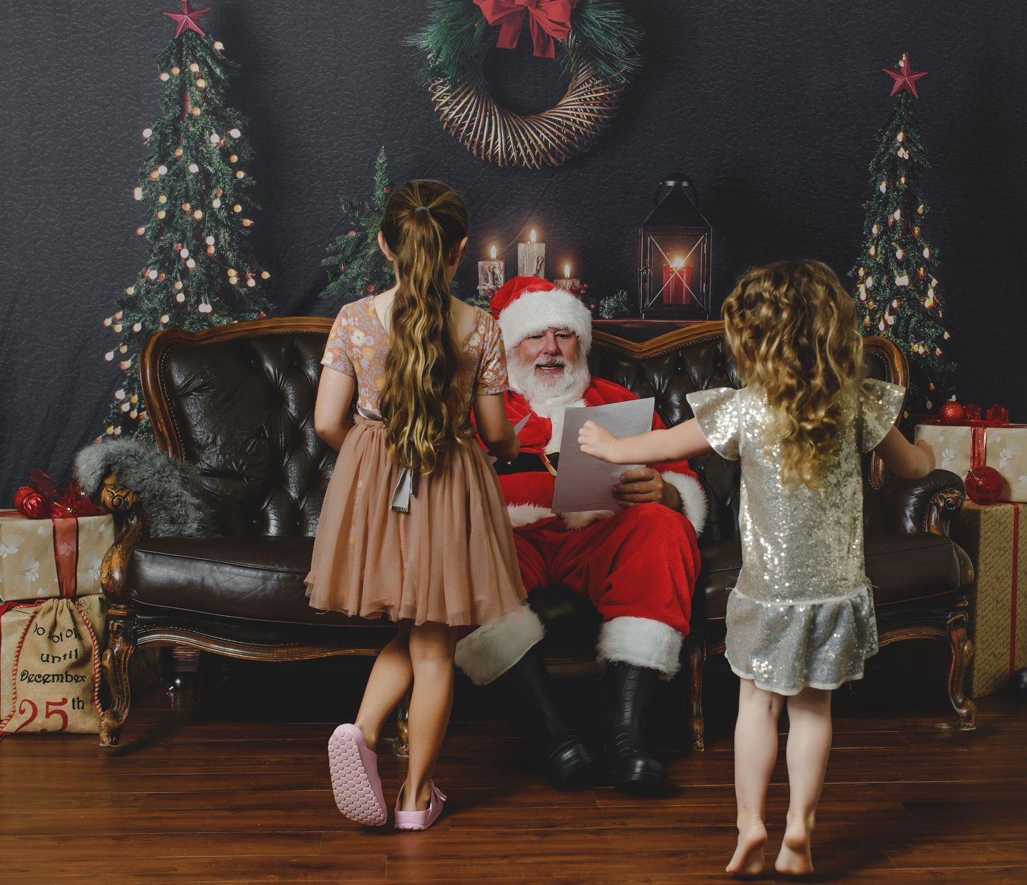 A holiday scene with Santa Claus seated on a leather couch between decorated Christmas trees and festive decorations.