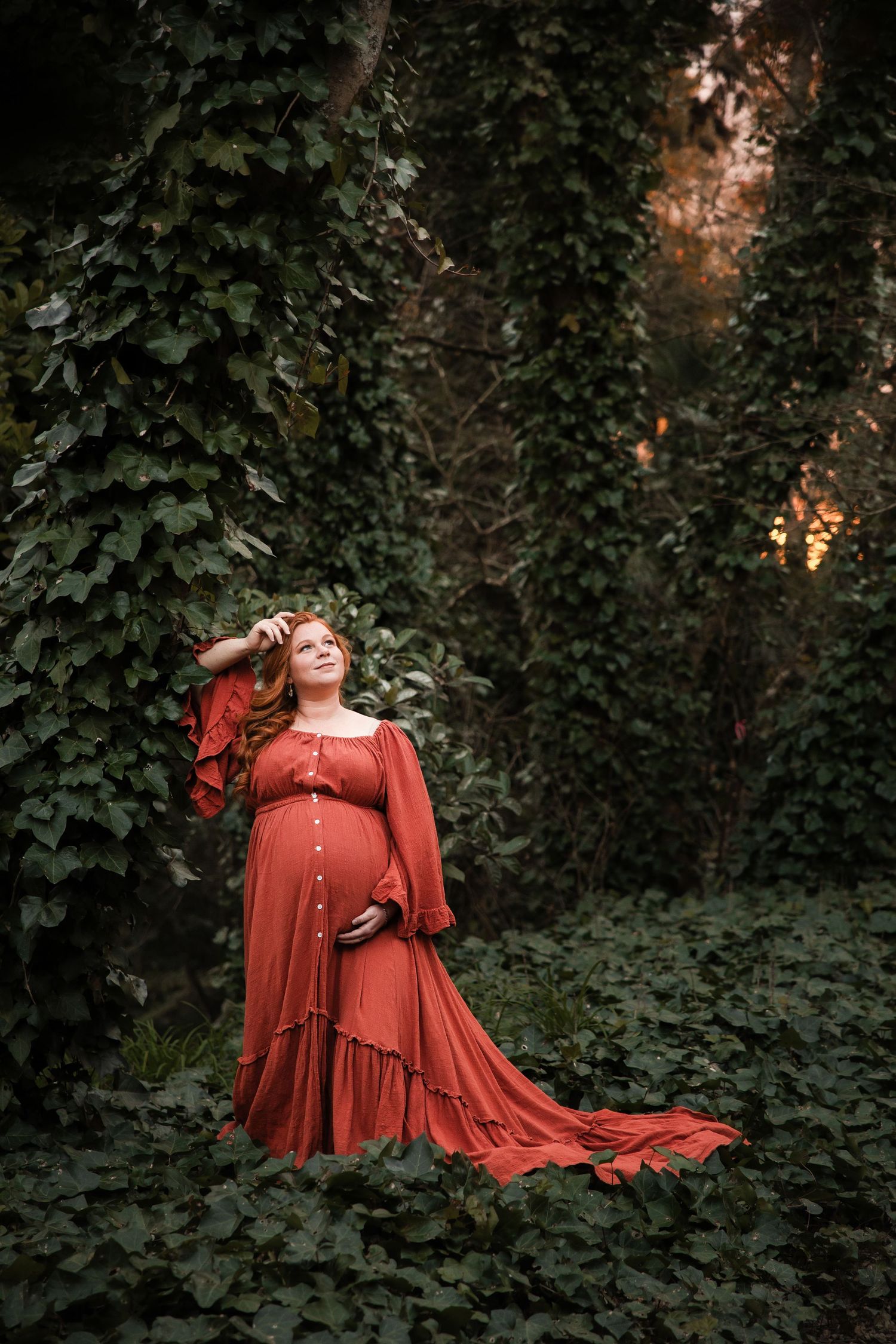 A figure in a flowing rust-red dress poses dramatically against a backdrop of lush green foliage at sunset.
