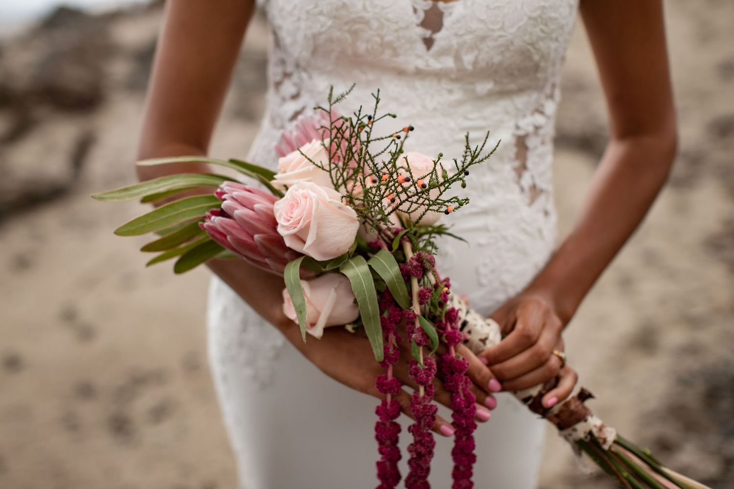 Close up of white lace wedding dress with pink and purple floral bouquet held against blurred rocky background.