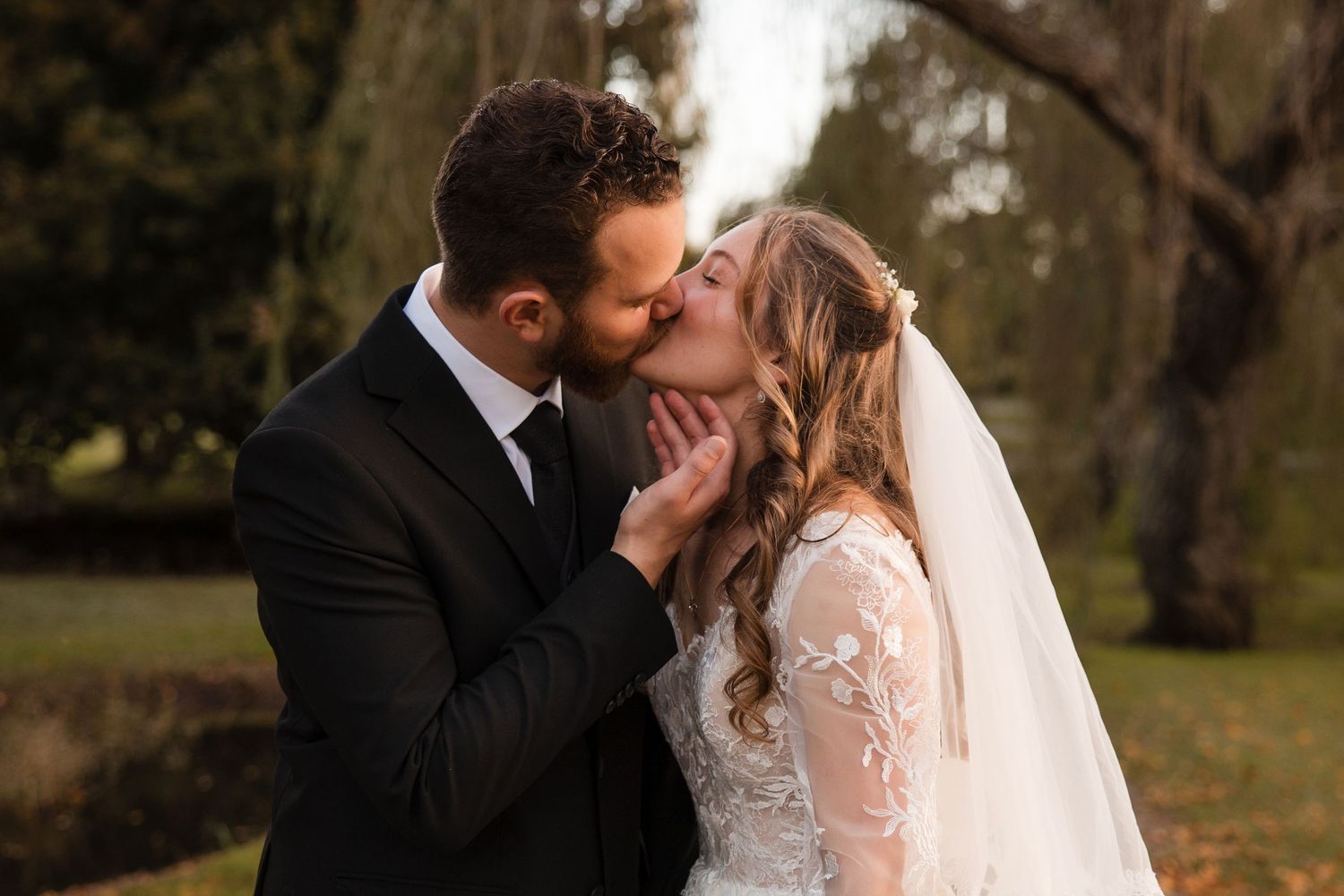 A bride in lace veil and groom in black suit share an intimate kiss during their outdoor autumn wedding ceremony.