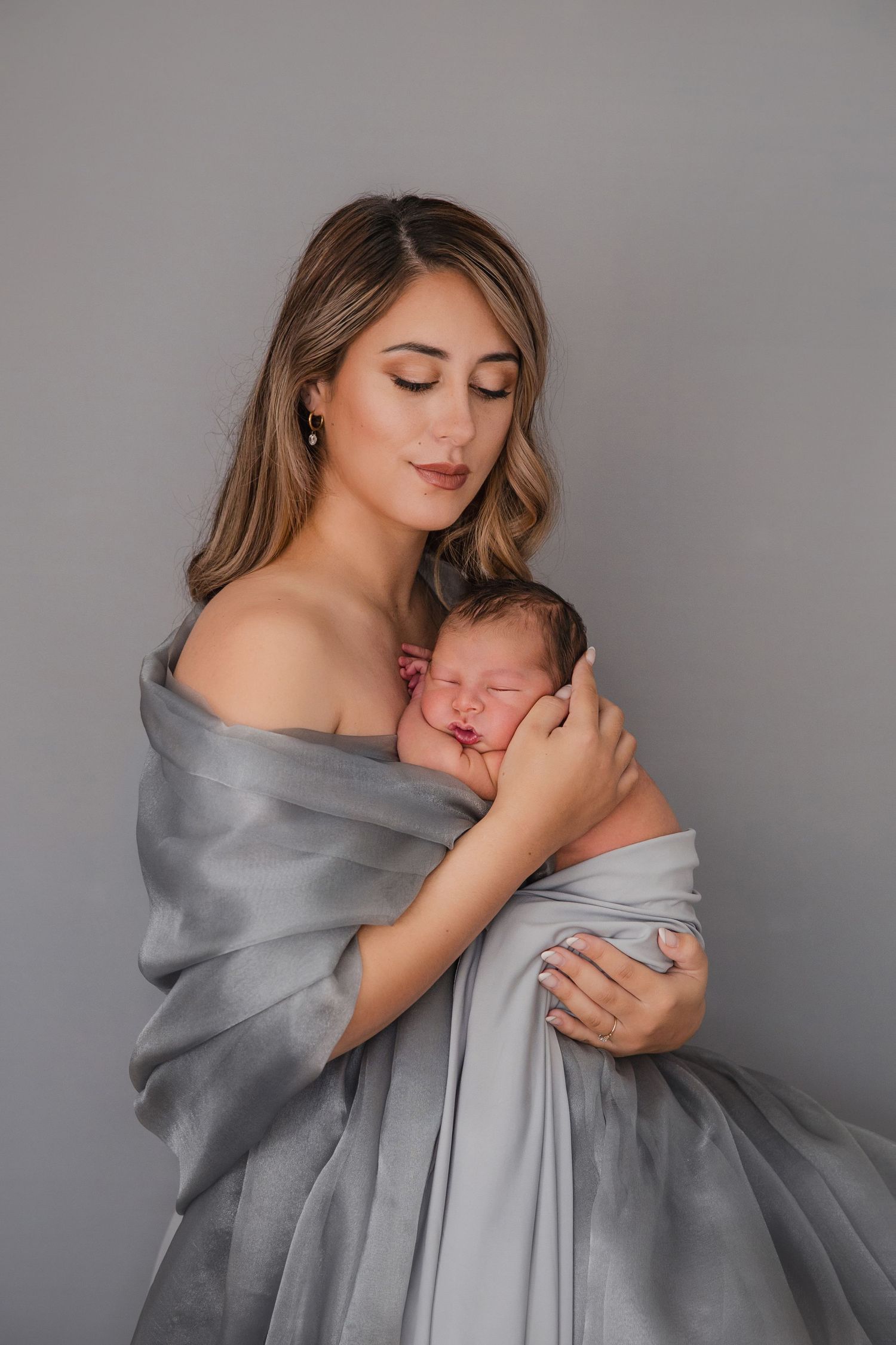 A young mother wrapped in gray silk fabric tenderly holds her sleeping newborn baby close to her chest.