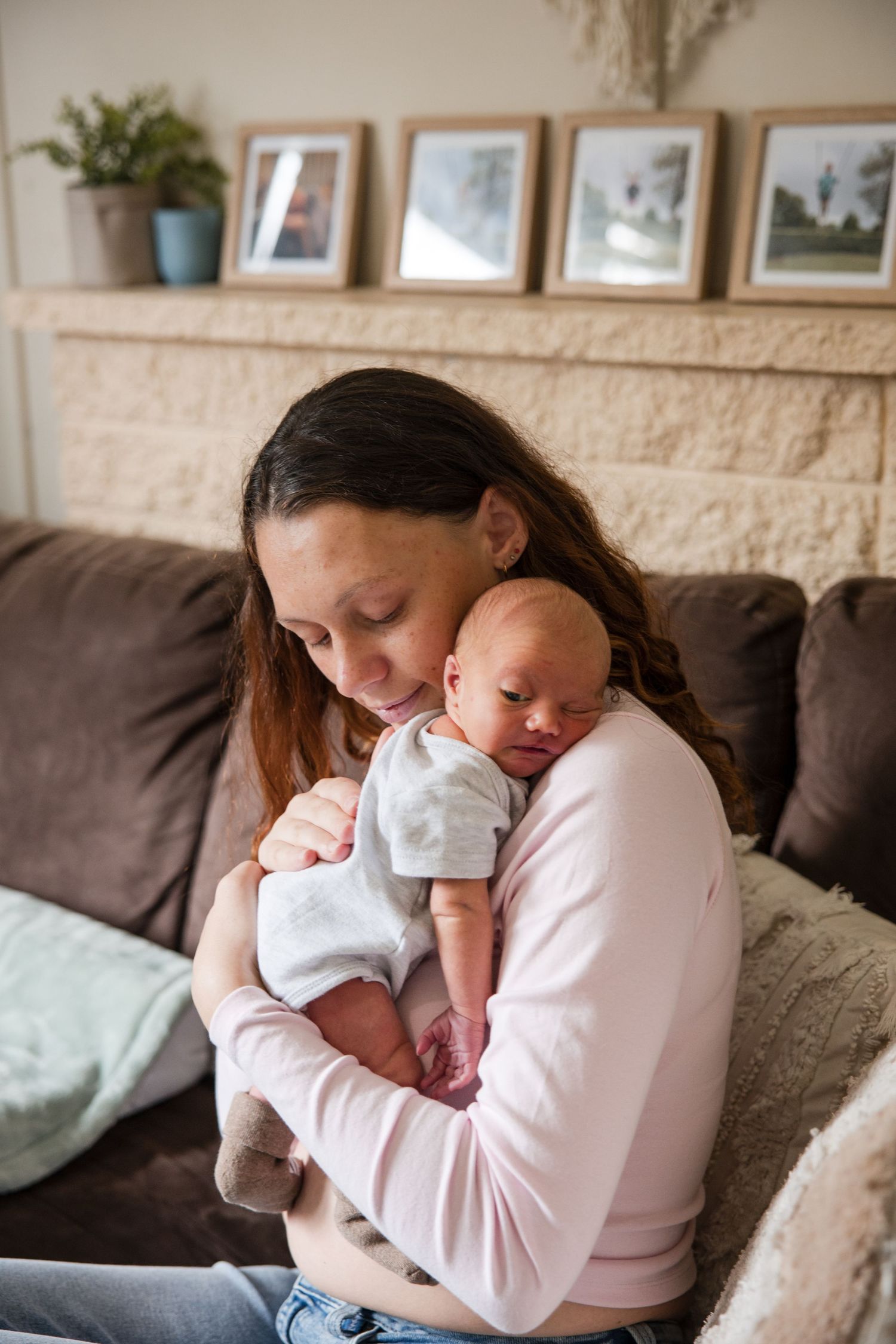 A mother tenderly holds her newborn baby close to her chest while sitting on a couch at home.