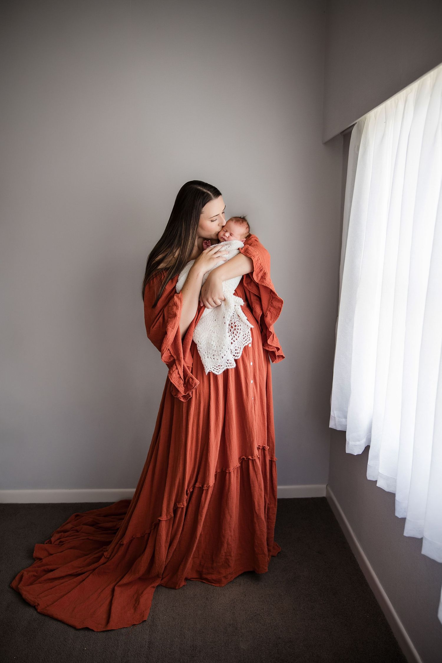 Mother in rust dress holds newborn baby near window in soft natural light.
