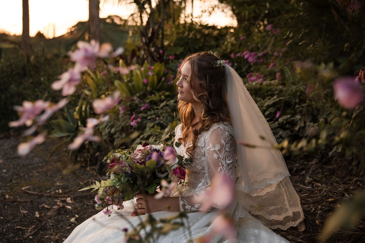 Soft pink flowers bloom in a garden setting at golden hour while someone sits in a flowy dress in the background.