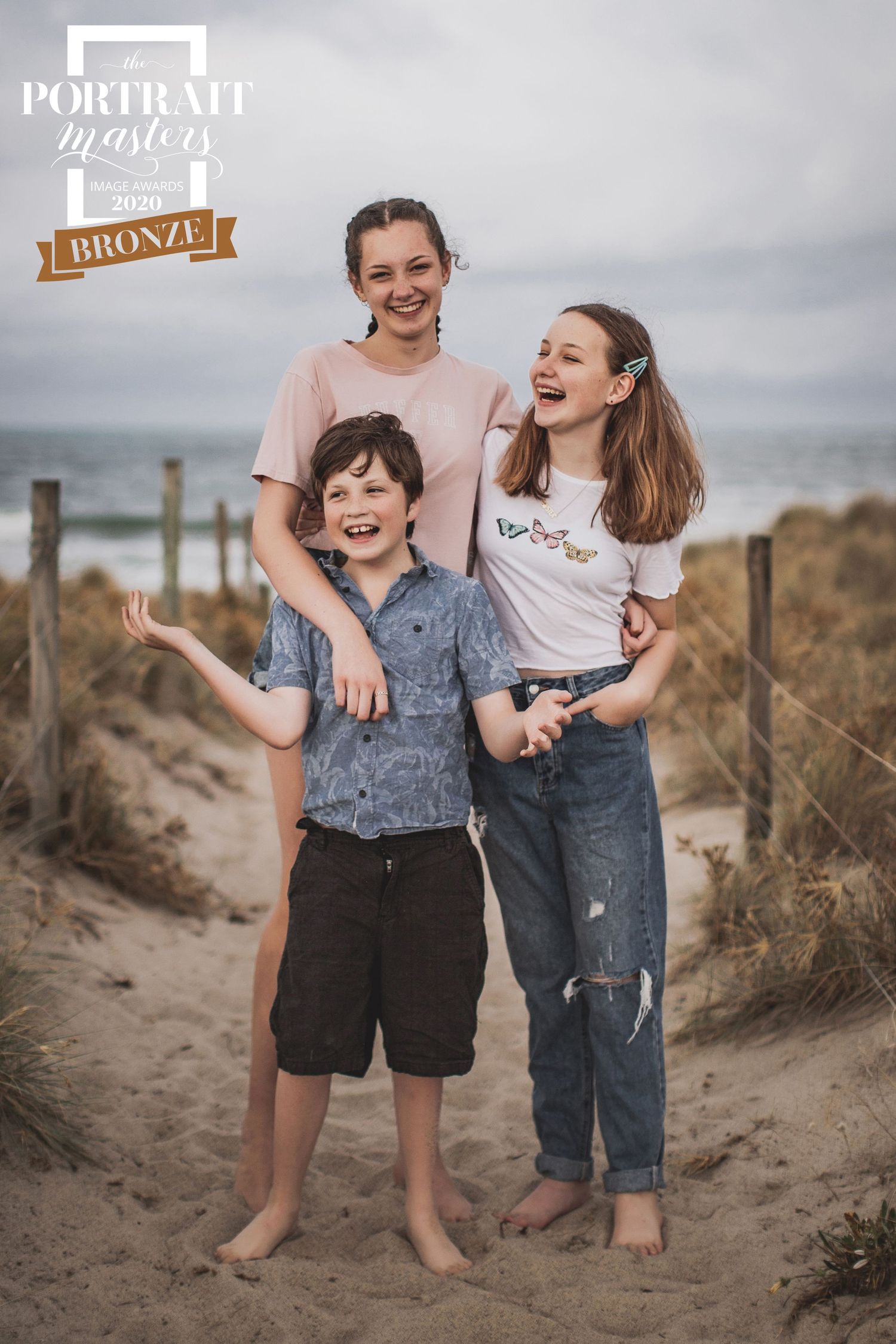 Three children laughing together on sandy beach path during overcast day, wearing casual clothing.