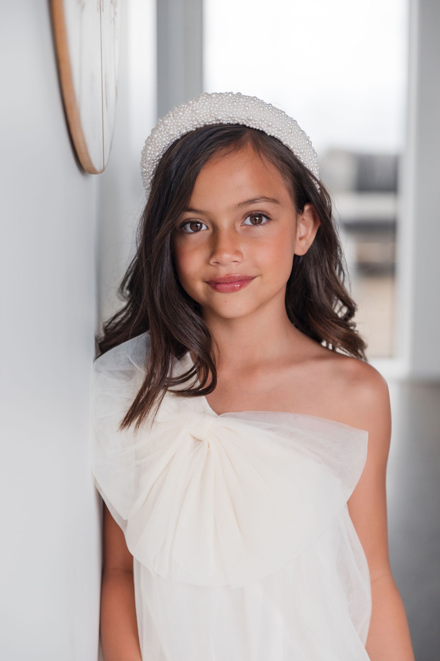Young person wearing a pearl headband and white off-shoulder dress smiles softly in an indoor portrait.