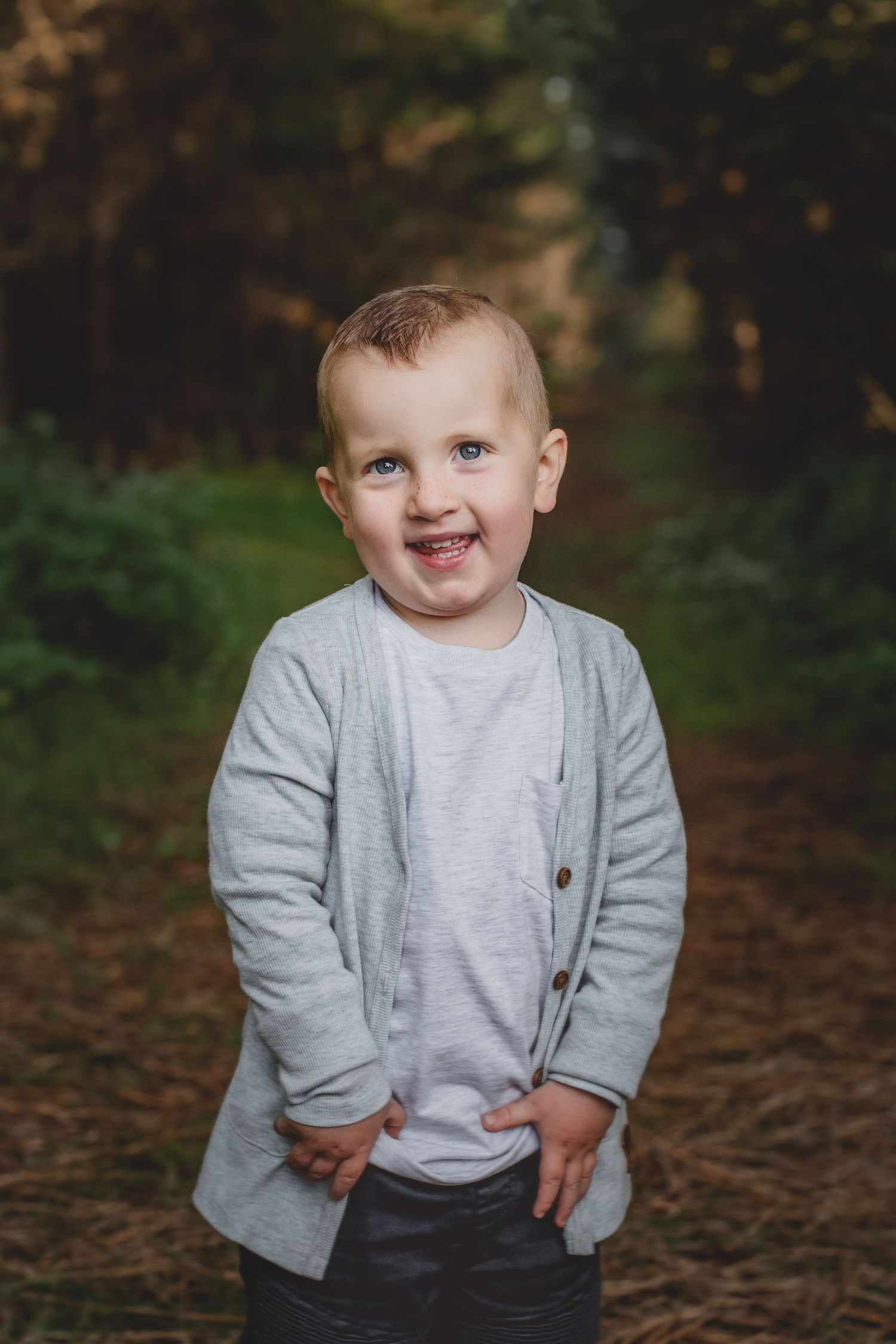 Young child in grey cardigan smiles while standing on forest path.