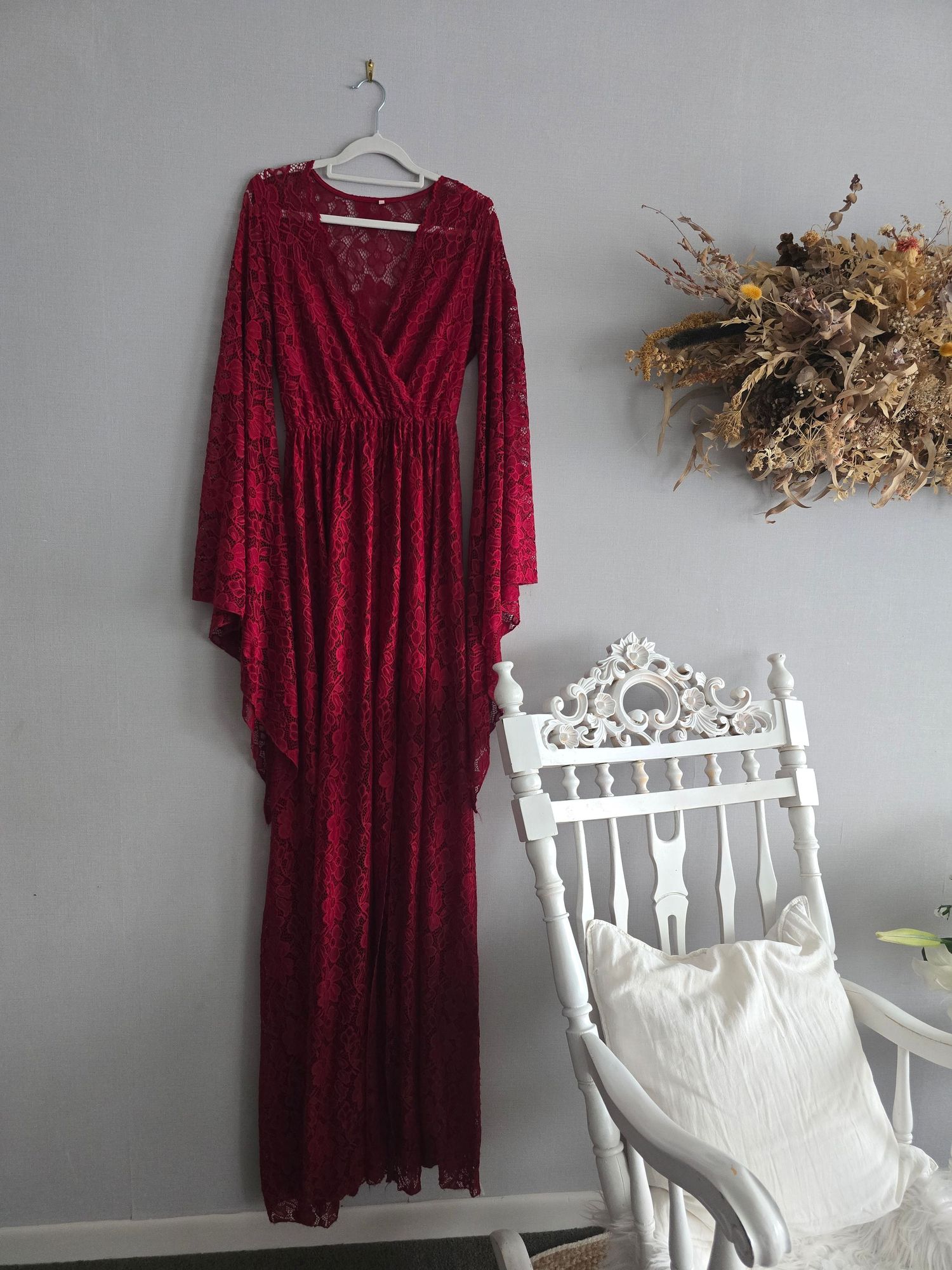 Red lace maxi dress with bell sleeves on hanger, displayed next to white ornate chair with fall floral decor.