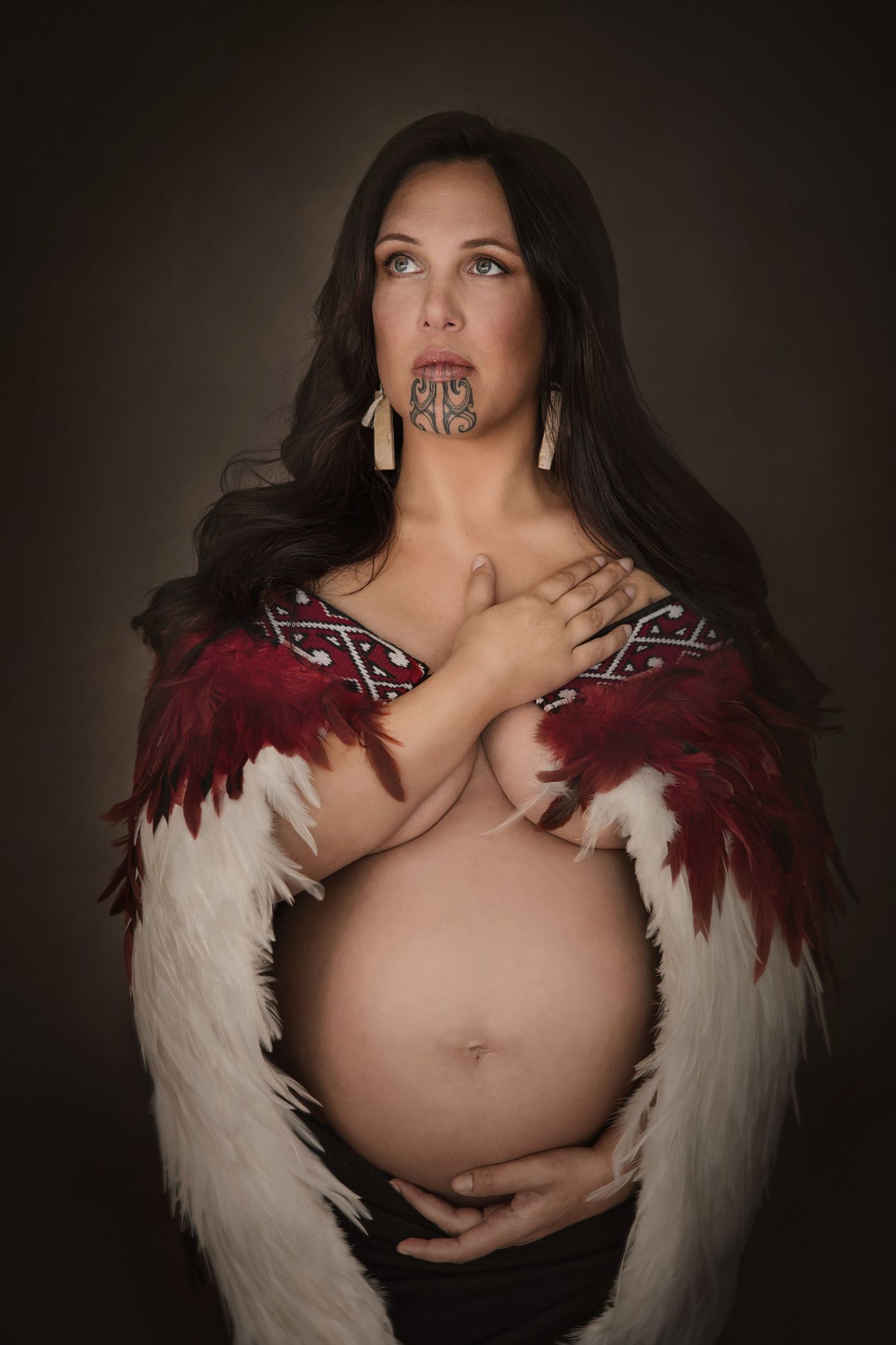 Pregnant Maori woman with traditional ta moko facial tattoo wearing red feather cloak, cradling baby bump in studio portrait.