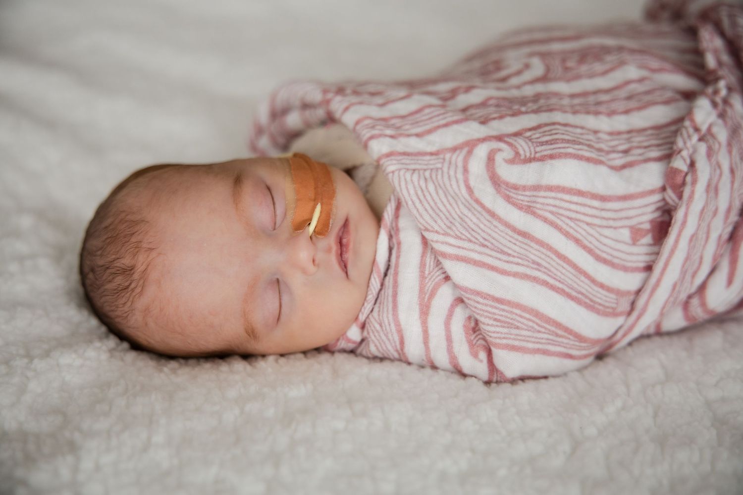 Swaddled newborn baby sleeping with nasal tube in pink striped blanket on white fur.