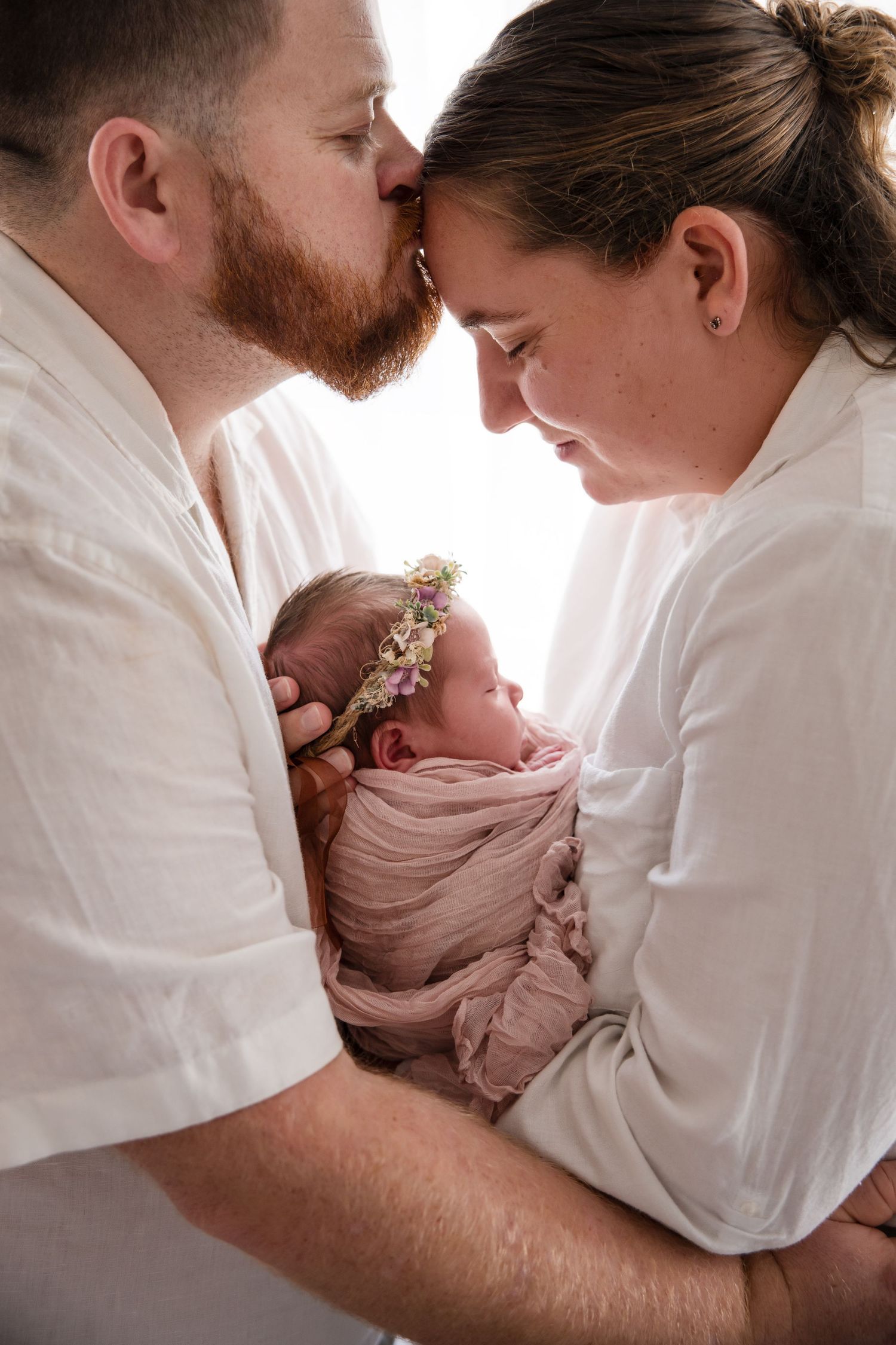 Parents in white shirts share tender moments with their newborn baby wrapped in pink swaddle and floral headband.