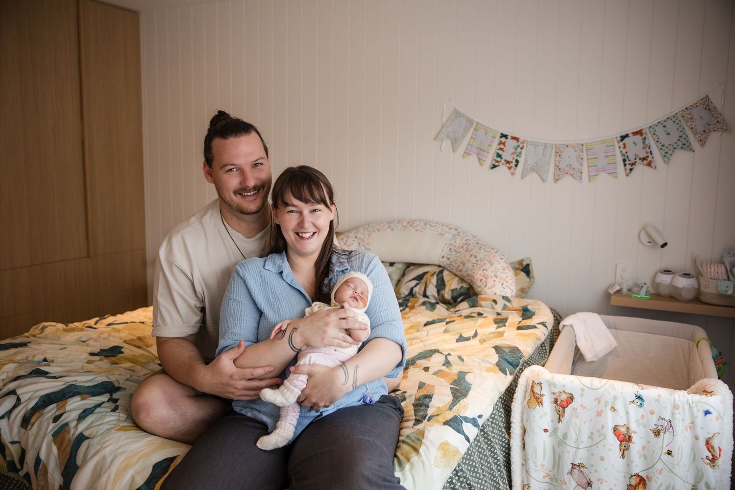 Happy couple sitting on bed holding newborn baby in cozy decorated nursery bedroom.