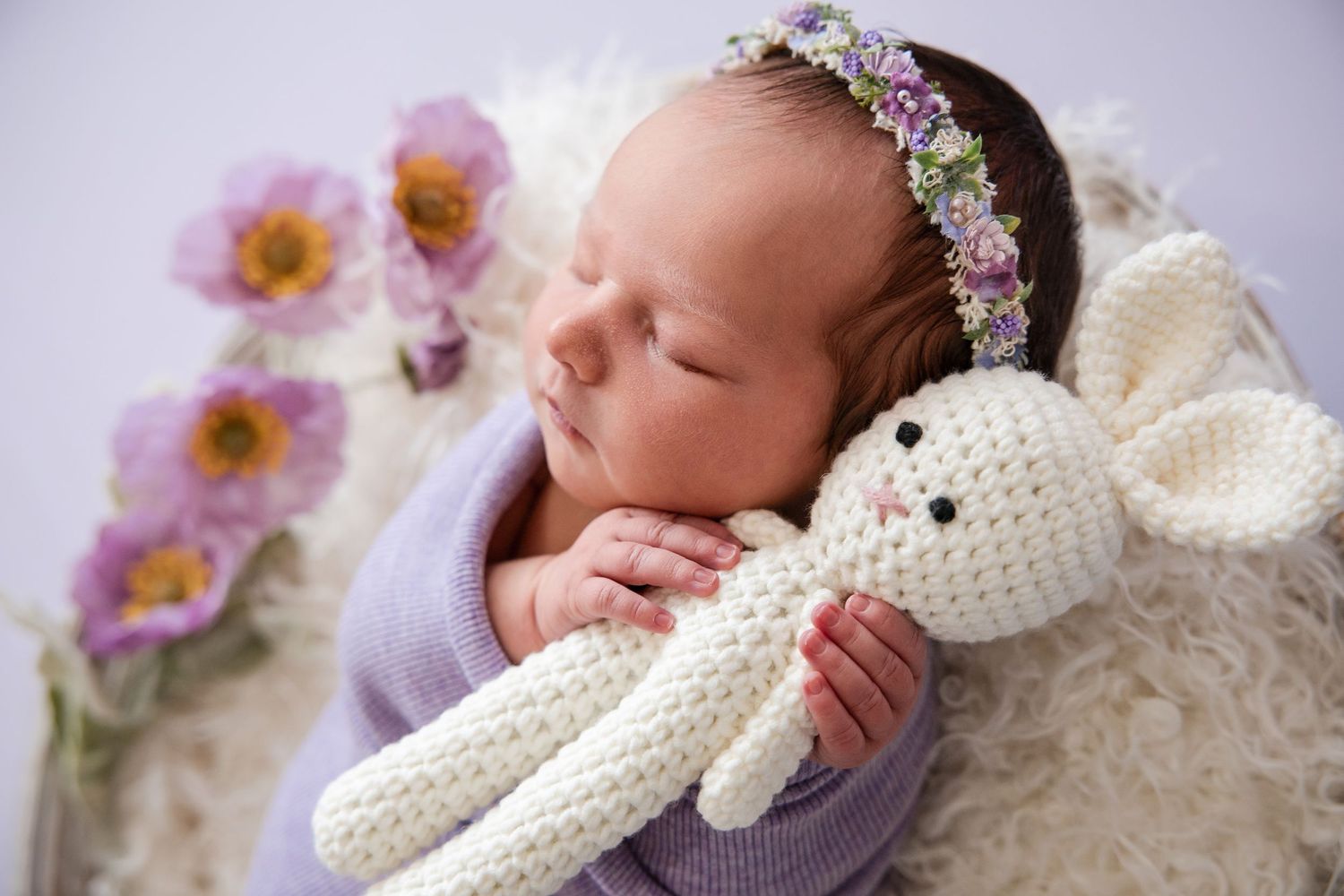 Newborn baby sleeping peacefully with white crocheted bunny toy and pink flowers in soft lighting.