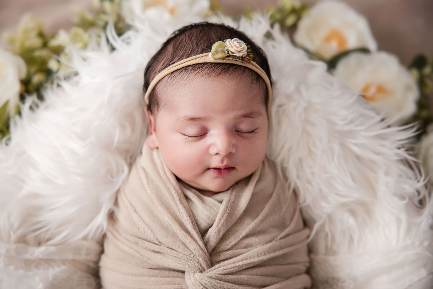 Peaceful newborn baby girl swaddled in beige wrap, wearing gold floral headband, sleeping on white fur with flowers.
