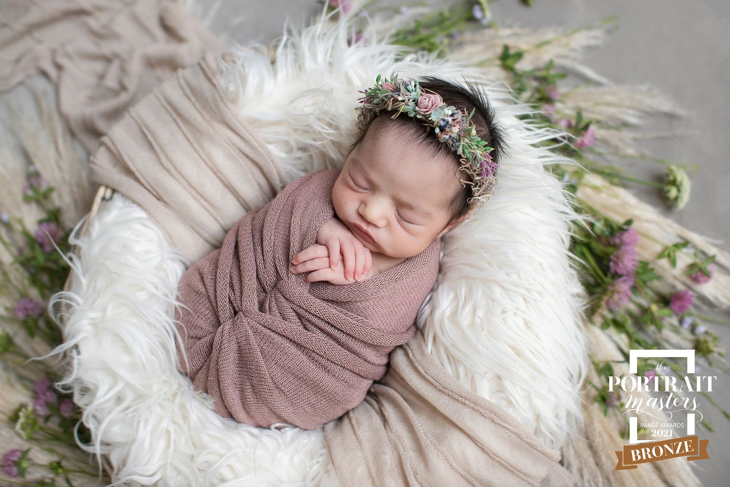 A newborn sleeping peacefully on a white fluffy blanket wearing a mauve wrap and delicate floral crown.