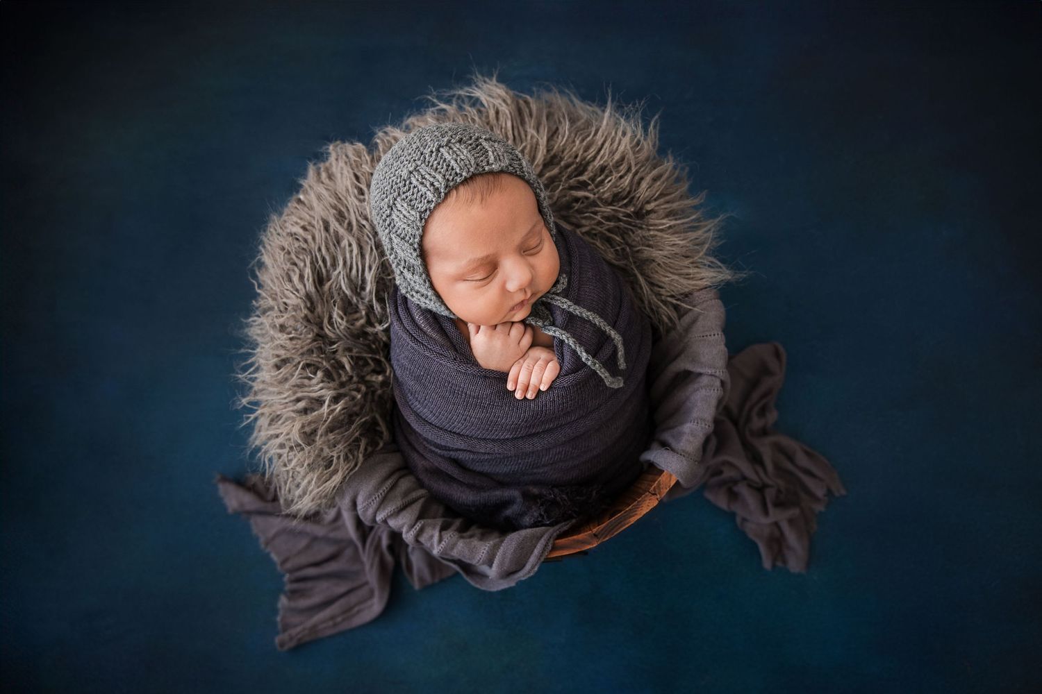 Sleeping infant wrapped in dark gray swaddle rests on furry blanket against navy background.