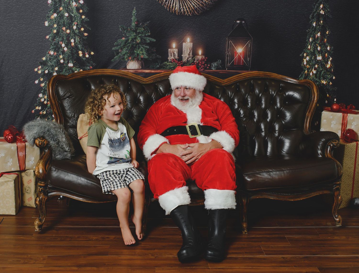 A child and Santa Claus share a moment on a vintage leather sofa with decorated Christmas trees and presents in the background.