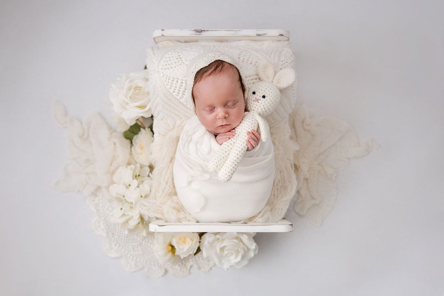 Newborn baby swaddled in white wrap holding a knit toy bear, nestled in a white wooden bed with cream flowers.