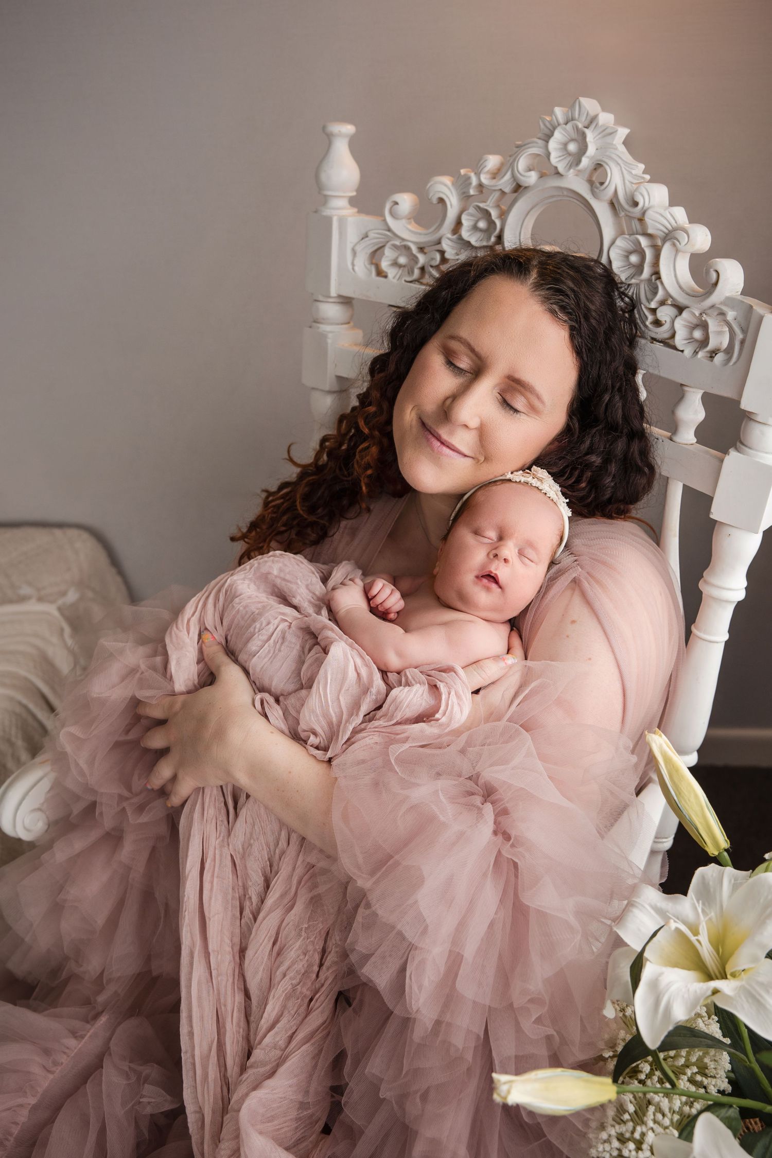 A newborn sleeping peacefully on a white ornate bench wearing a pink tulle dress with white lilies in the scene.