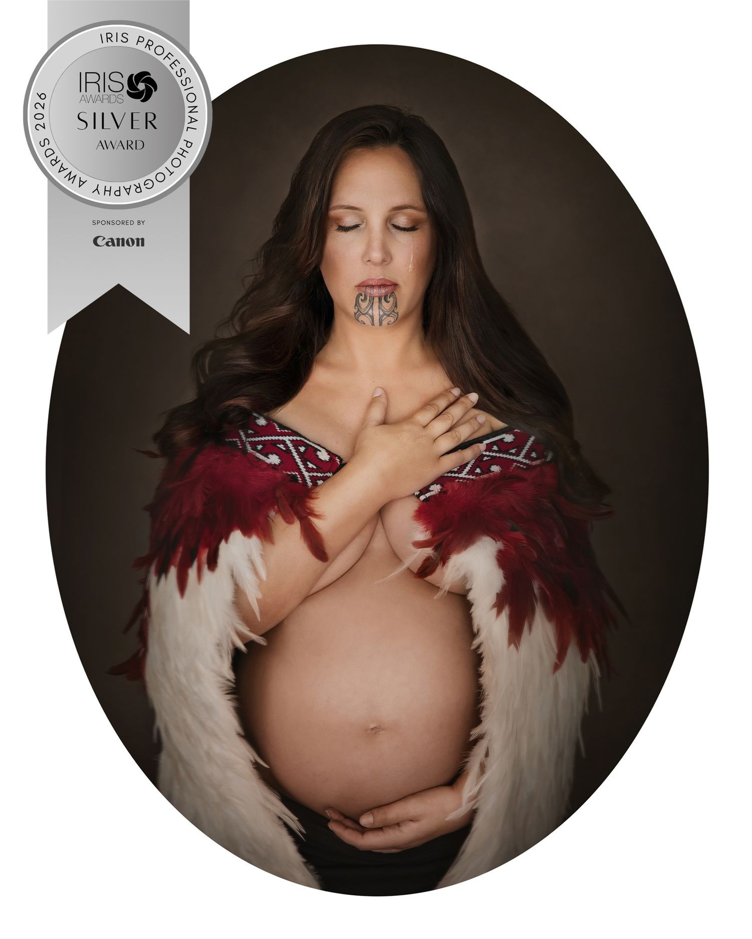 Award-winning maternity portrait of a Māori woman with tā moko, wearing a traditional feathered cloak, cradling her pregnant belly.