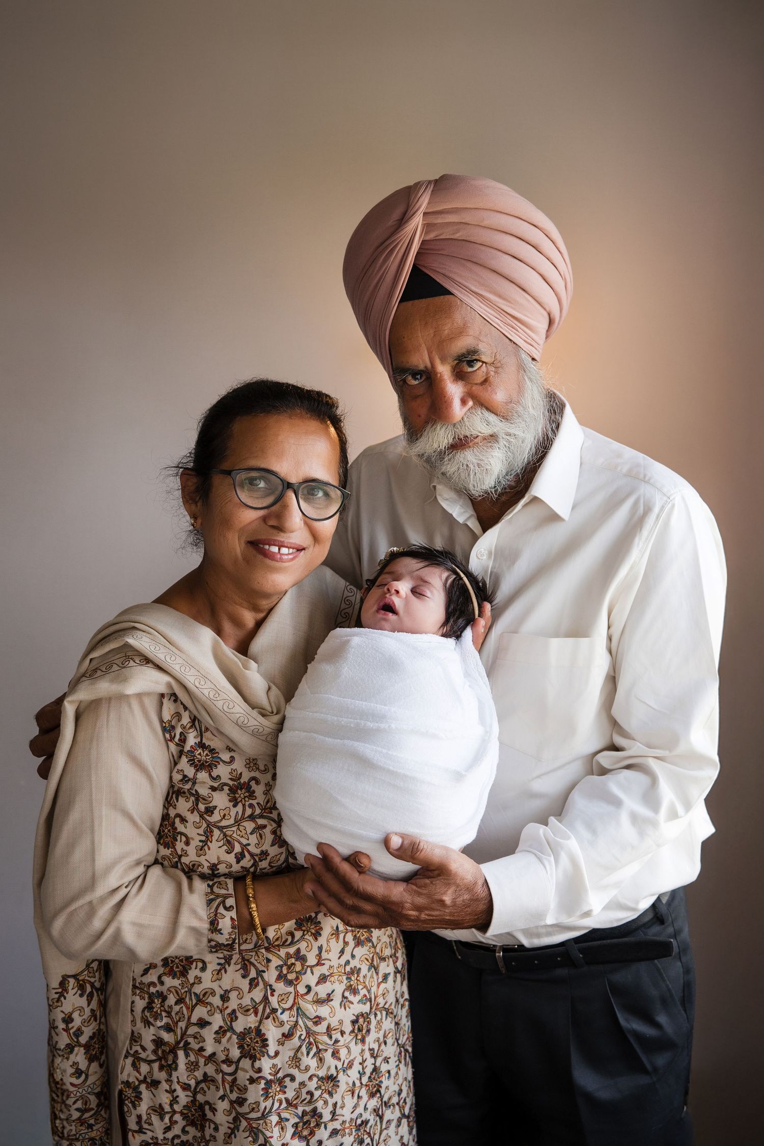 Elderly Sikh couple in traditional attire smiling while holding a swaddled newborn baby.
