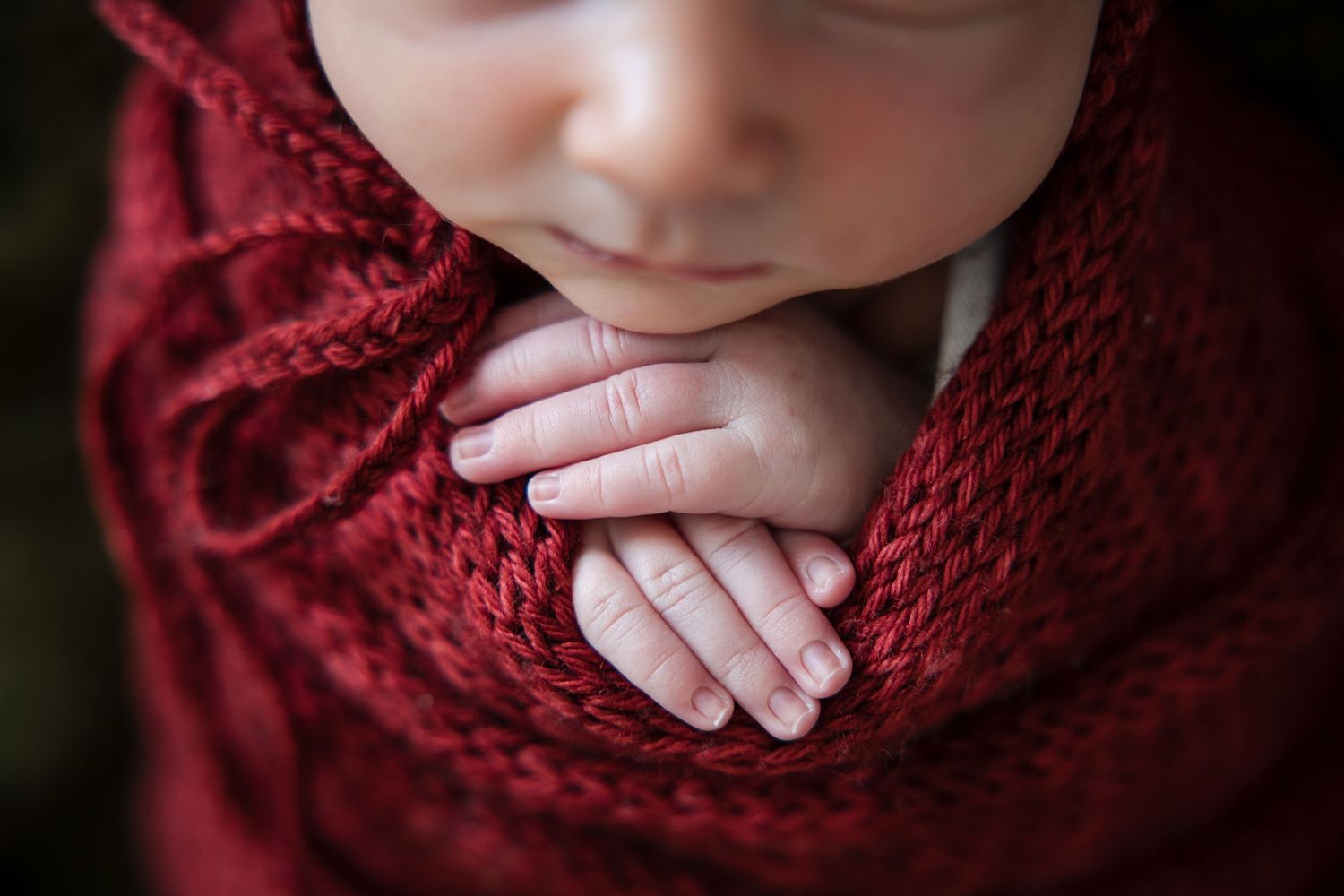 Close-up of a sleeping newborn baby wrapped in a soft red knit blanket with tiny hands tucked under chin.