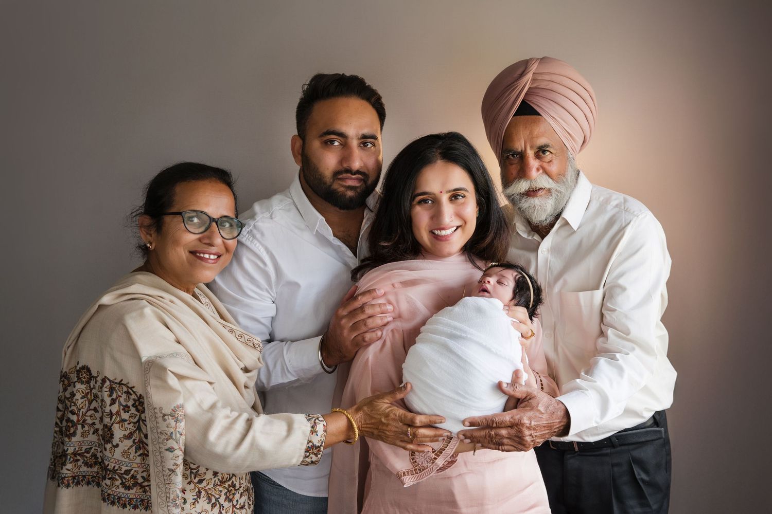 Sikh family portrait with newborn baby, grandparents and parents smiling together in a studio setting.
