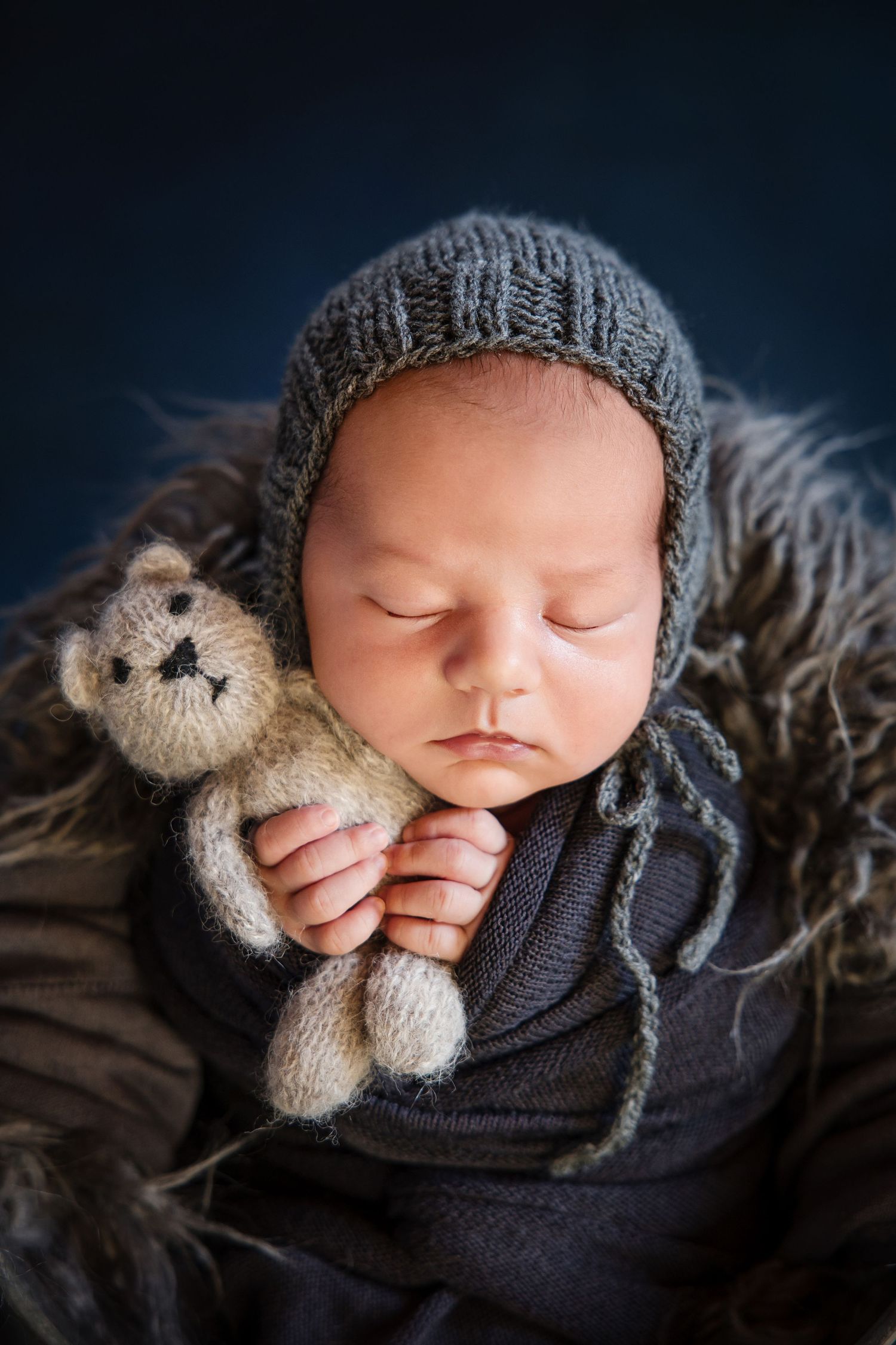 Sleeping newborn baby wearing a gray knit bonnet, wrapped in navy fabric, holding a small teddy bear on a fur blanket.