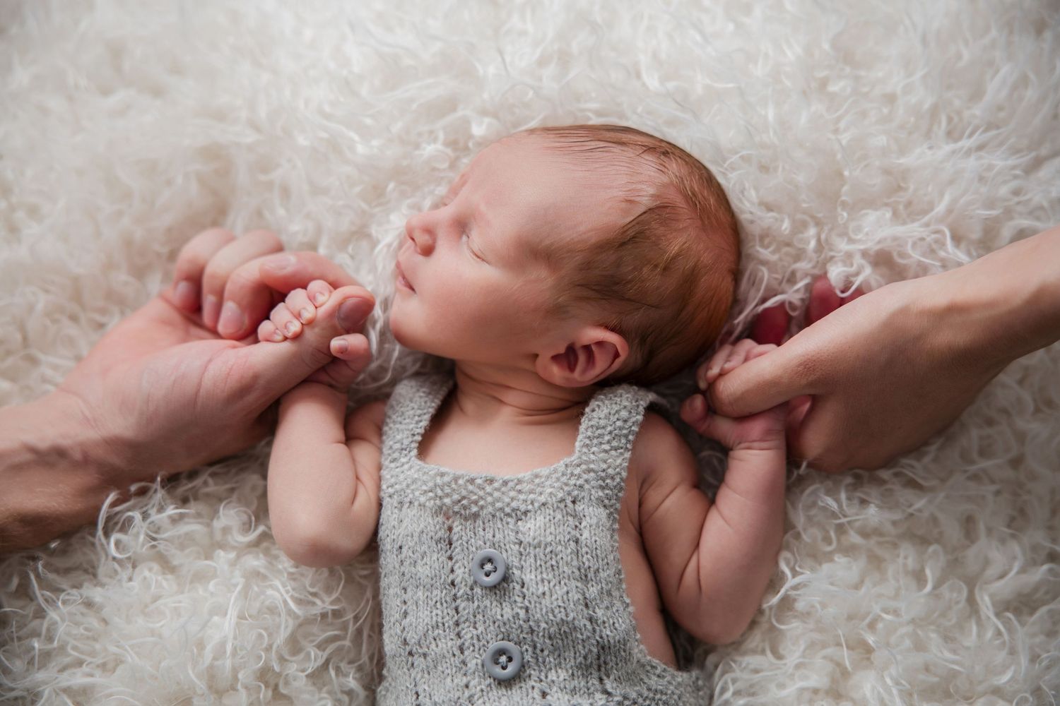 Newborn baby wearing gray knit clothing lies on white lace blanket while adult hands gently touch their face.