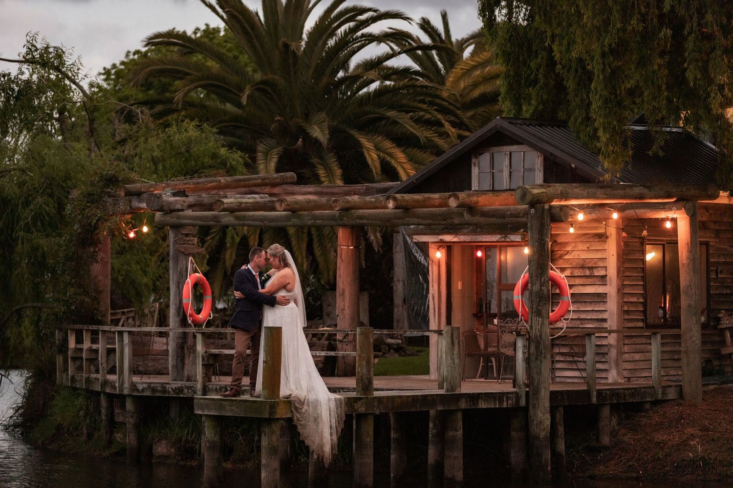 A romantic sunset scene on a rustic wooden dock decorated with string lights and life preservers amid palm trees and water.