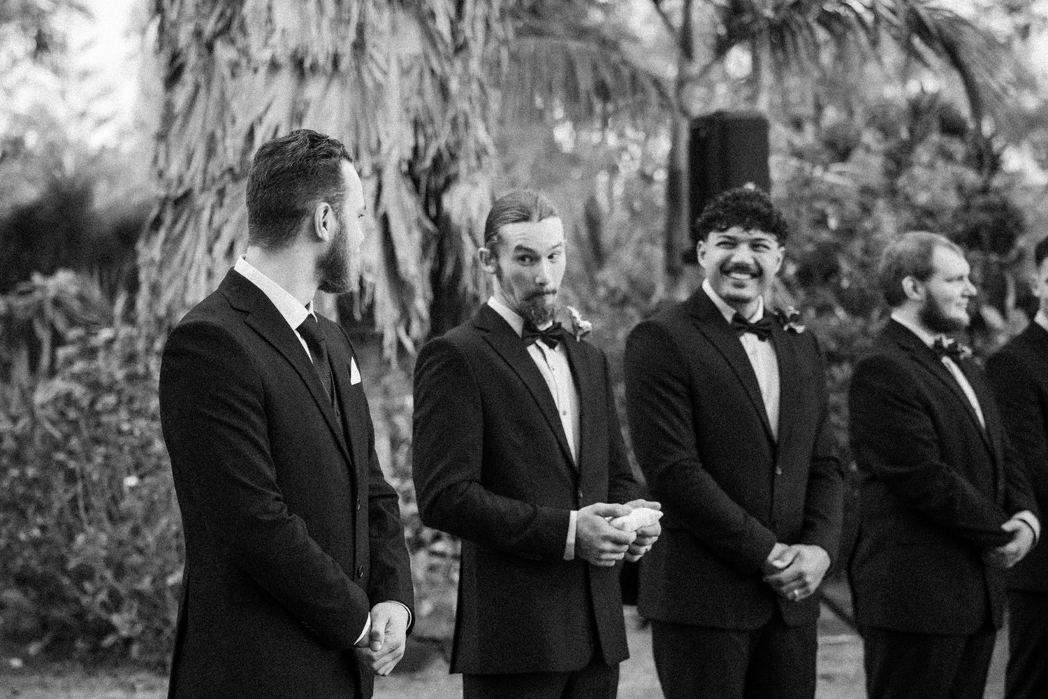A black and white photograph shows groomsmen in tuxedos standing together outdoors with lush foliage in the background.