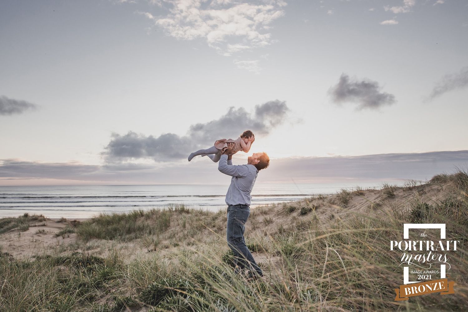 A person lifts a child against a dramatic sunset sky on a grassy coastal dune, with ocean visible in the background.