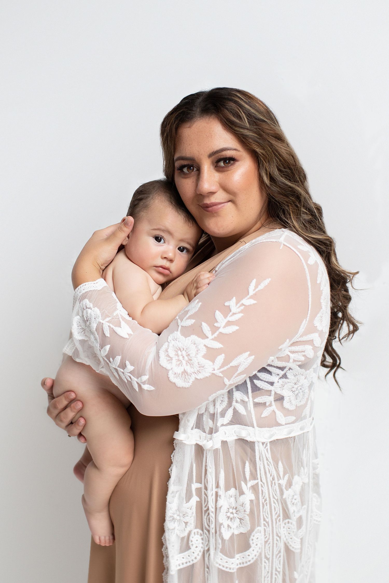 A mother in white lace robe tenderly holds her newborn baby against a soft white background.