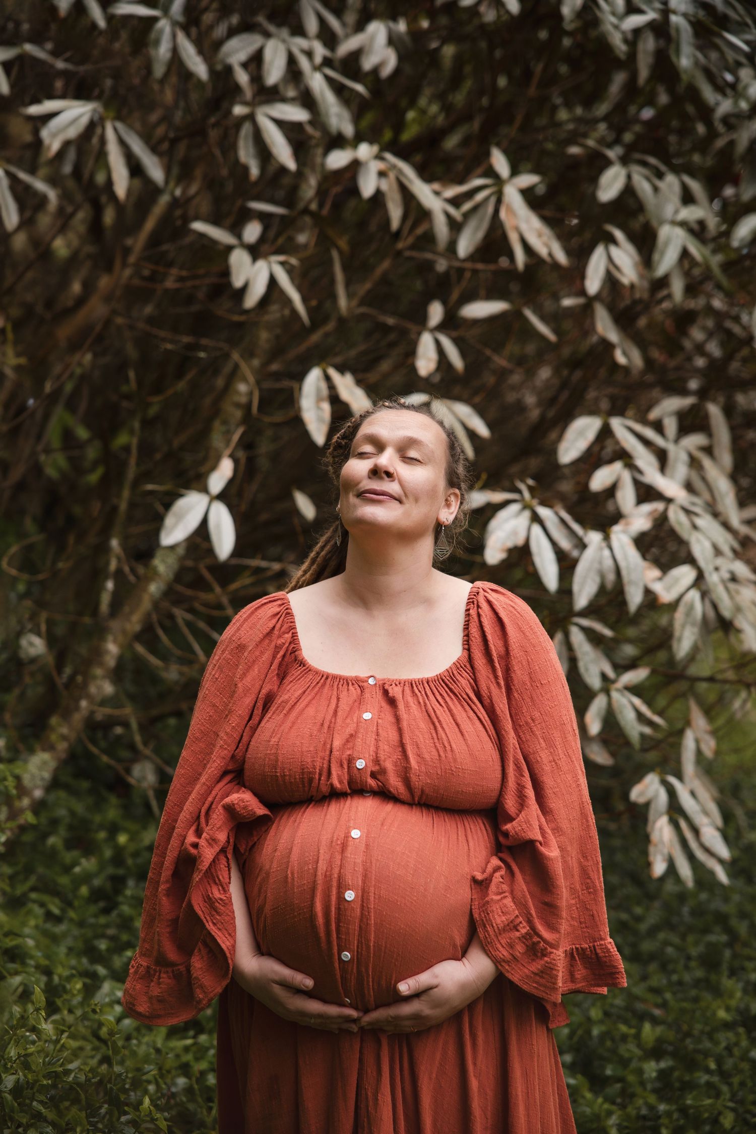 A pregnant person in a rust-colored dress stands peacefully with eyes closed beneath white flowering branches outdoors.